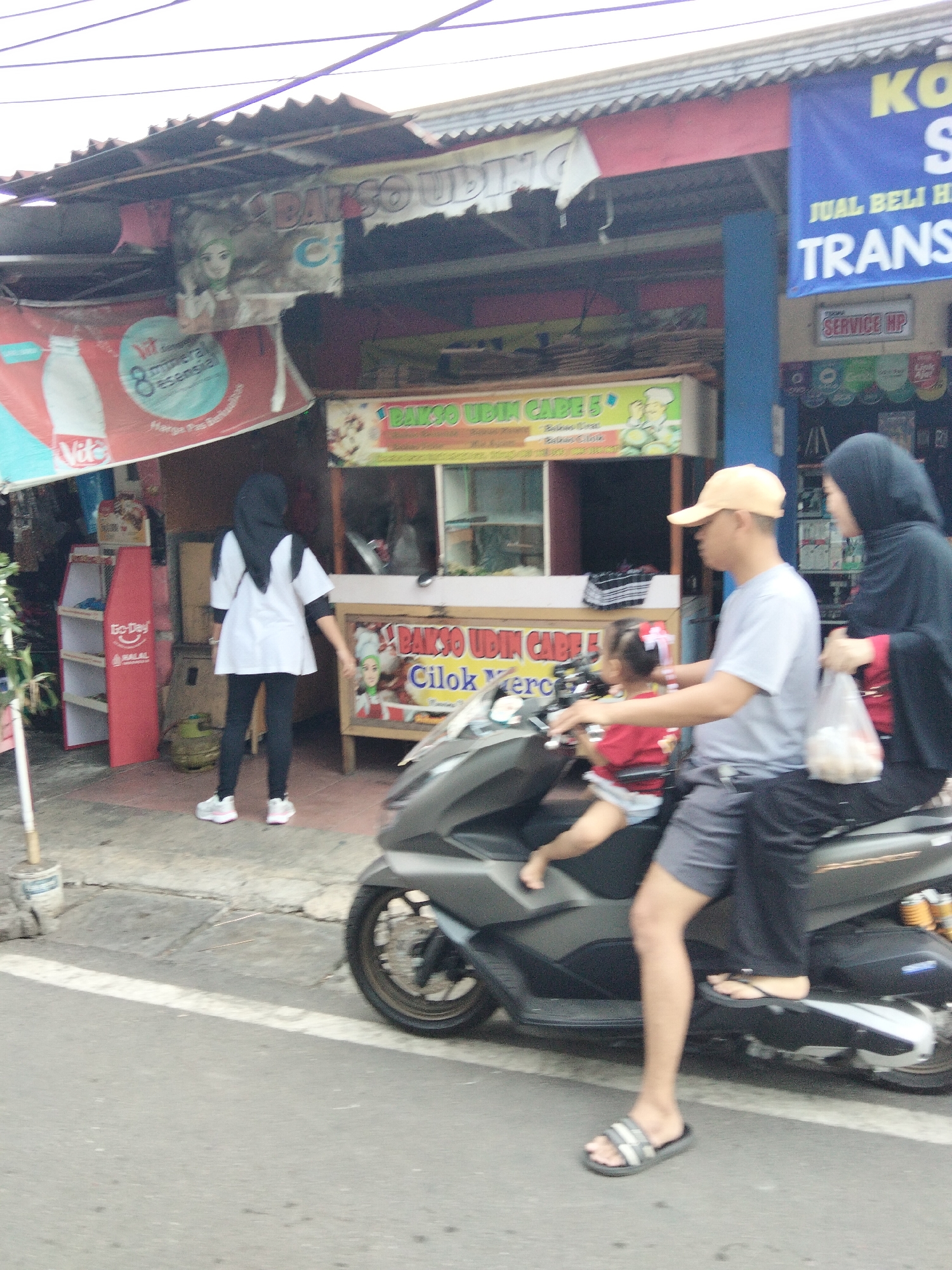 The image depicts a street scene in South Tangerang, Indonesia, characterized by a modest storefront and a busy roadside. In the foreground, a scooter is prominently featured, with a man in a light grey t-shirt and dark shorts, wearing a tan baseball cap, operating it. He is accompanied by a young child in a red shirt, seated in front of him. Behind them, a woman wearing a dark hijab and a red top is seated on the scooter, holding a white plastic bag filled with items.
The background reveals a street-side food stall, likely selling "Bakso Udin Cable" as indicated by signage. Various banners and advertisements are visible on the building, including one with a red and white design featuring a bottle and text that appears to be promotional. To the left of the stall, a woman dressed in a white tunic and black leggings, with a black hijab, stands on the sidewalk. Next to her is a red shelf unit displaying some products, and a green gas cylinder is visible on the ground. The street itself is paved with asphalt and marked with a white line. The overall environment suggests a bustling urban setting with commercial activity.