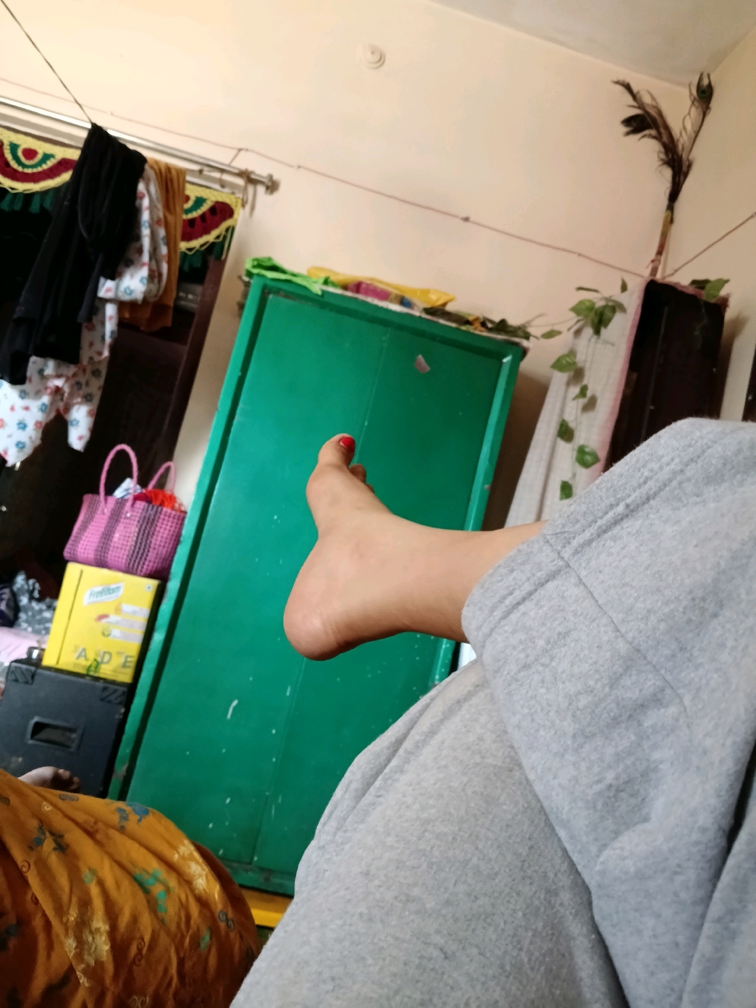 The image depicts an indoor scene in what appears to be a residential setting in C Camp, India. The focus is on a person's bare foot, which is resting on light gray sweatpants. The foot has red nail polish on the toenail. In the background, a vibrant green cabinet is visible, along with a pink woven bag and a yellow box stacked on top of a black case. Clothes are hanging on a rod on the left side of the frame, and a peacock feather is displayed on the right. The walls are a neutral, light color, and a vine with green leaves trails down from the top right. The overall impression is that of a casual, domestic environment.