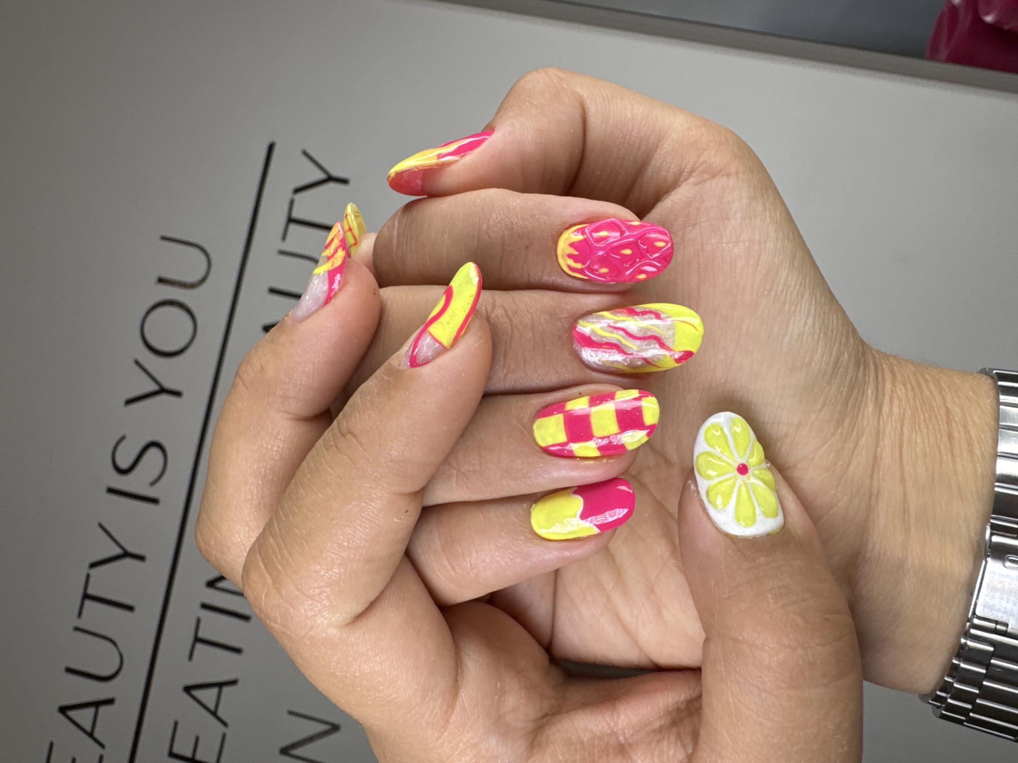 A close-up depicts a person's hand showcasing brightly colored nail art. The nails are designed with a vibrant color palette of yellow and pink and feature different patterns: abstract waves, geometric grids, and one nail has a design resembling a lemon. The person wears a stainless steel wristwatch. The hand rests on a gray surface printed with the text "BEAUTY IS YOU." The location is Podgorica, Montenegro.