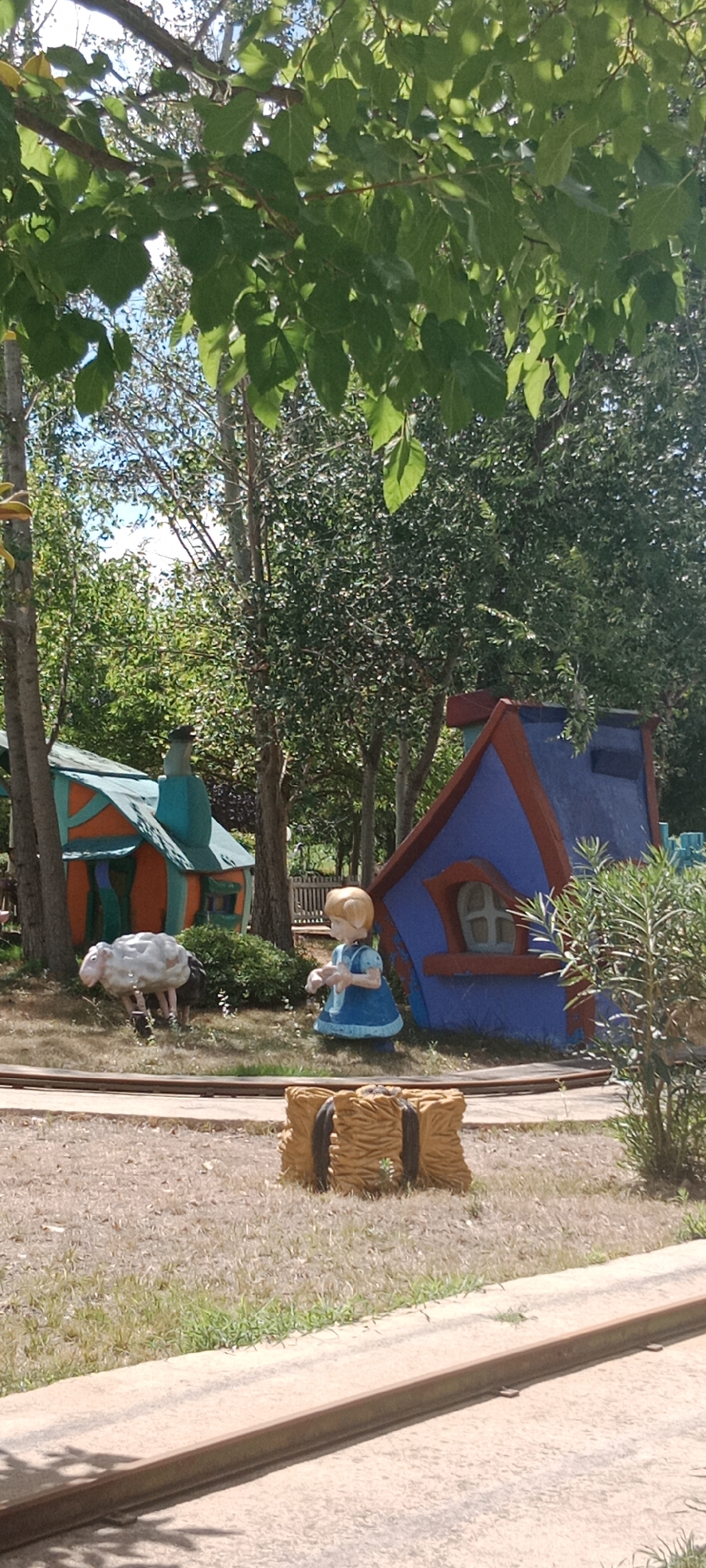 The image captures a scene likely within a theme park or amusement area. In the foreground, a set of train tracks runs horizontally across the frame. To the left of the tracks, a white, stylized sheep figure is positioned on the dry, grassy ground. Further to the right, a statue of a young girl in a blue dress stands facing the sheep. Behind the figures, two small, whimsical structures are visible. The structure on the left is a house painted in orange and green, with a conical blue roof section. The structure on the right is a taller, blue house with red trim, featuring a small, square window. The background is filled with mature trees with dense green foliage, suggesting a natural, shaded environment. The overall impression is that of a playful, decorative display designed to evoke a storybook or fairy tale setting. The image does not contain any explicit markers or clues that would pinpoint its location within Torrejón de Ardoz, Spain.