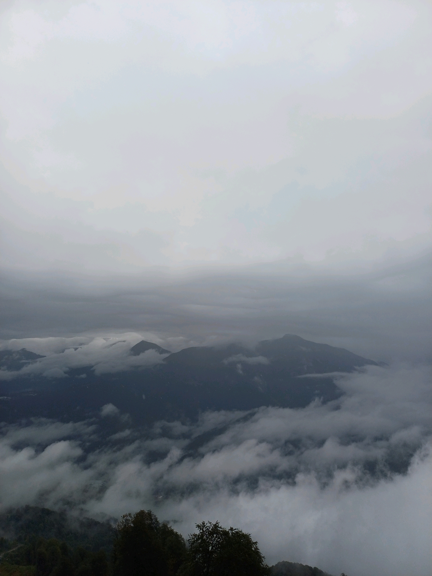 This image depicts a vast mountain landscape, likely taken from a high vantage point in Estosadok, Russia. The scene is dominated by a thick, overcast sky with layers of grey clouds obscuring the upper reaches of the mountains. Below the clouds, several mountain peaks emerge, some sharply defined and others softened by the pervasive mist. Wisps and banks of fog swirl around the mountains, creating a sense of depth and mystery. The lower foreground of the image shows a dark, forested slope with silhouetted trees, suggesting the photograph was taken during the late afternoon or on a heavily clouded day, as light is dim. There are no people, animals, or discernible man-made objects visible in the frame. The environment is exclusively natural, characterized by rugged terrain and atmospheric conditions.