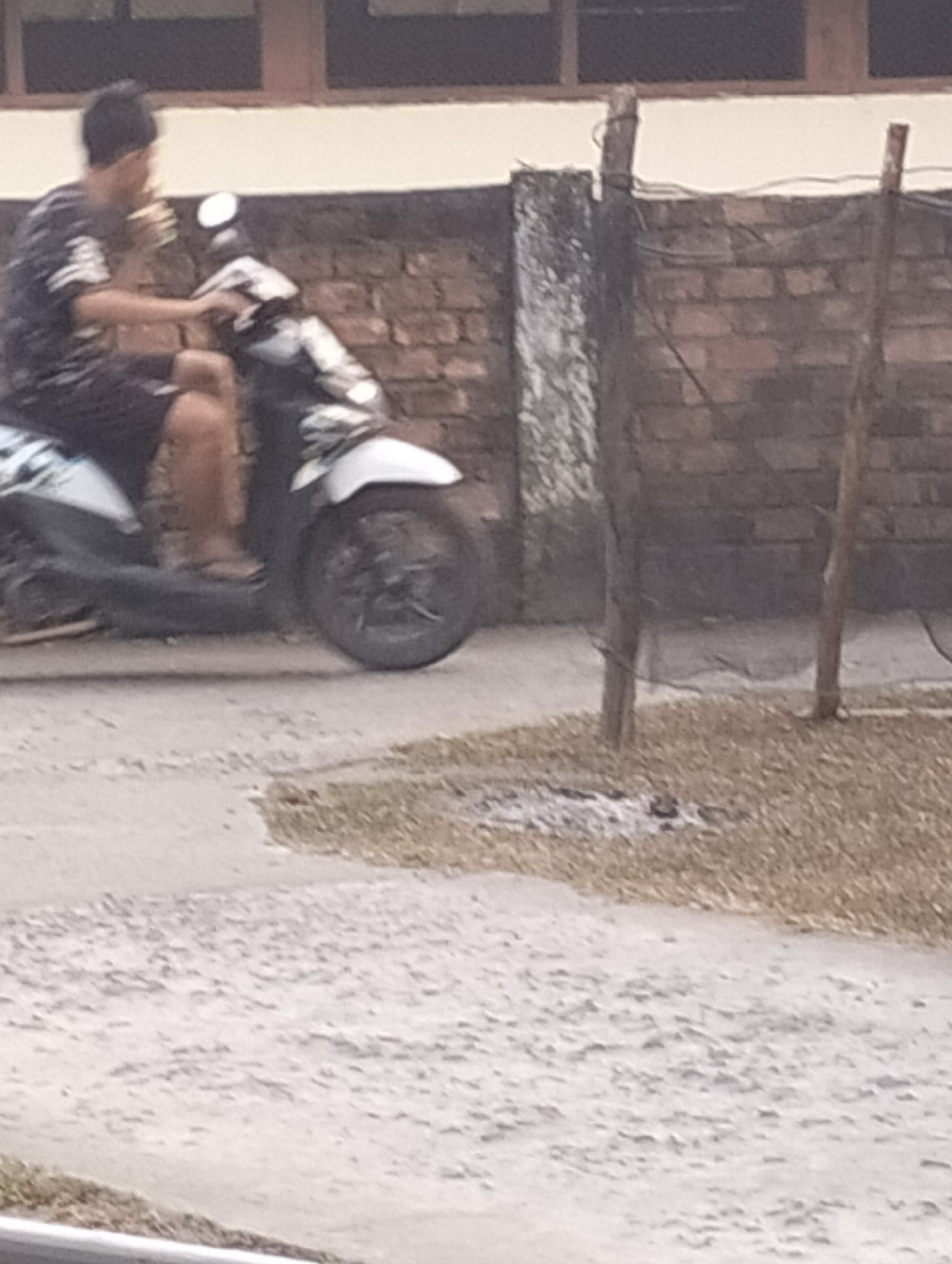 In Tanjung Laut, Indonesia, a person is riding a motorcycle. The rider, wearing a dark patterned shirt and dark shorts, is seated on the motorcycle and appears to be holding something up to their mouth, possibly eating. The motorcycle is dark and white, with a visible front wheel and part of the rear wheel. The setting appears to be outdoors, near a brick wall. The ground is paved with some dry grass and debris in the foreground. Wooden posts and what appears to be netting are present near the brick wall, suggesting a fence or boundary. The background shows a building with windows above the brick wall.
