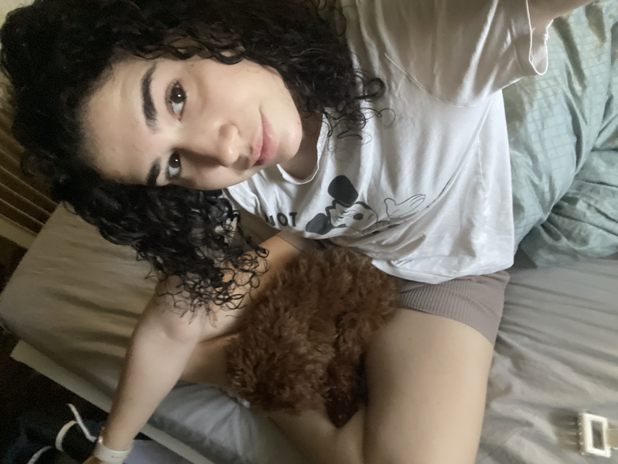 A young woman with dark curly hair is taking a selfie in what appears to be a bedroom. She is wearing a white t-shirt with a Mickey Mouse design and neutral-colored shorts. A small brown poodle or poodle mix is nestled on her lap. The bed is covered with light-colored sheets and a muted green comforter or duvet. The environment seems to be lit by natural light. There is no indication that this image was taken in Sofia, Bulgaria.