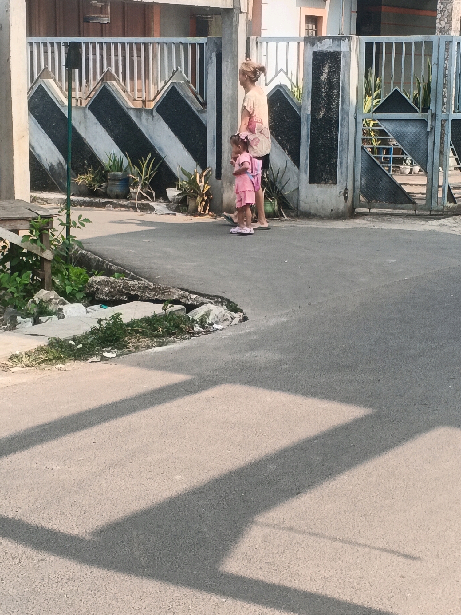 In Kadu, Indonesia, a woman and a young child stand on an asphalt street in front of a gated property. The woman, with blonde hair pulled back, wears a patterned t-shirt and shorts. The child, facing away from the camera, is dressed in a pink dress and sandals, carrying a pink backpack. Behind them, a metal gate with a diamond pattern and vertical bars is closed. The property gate features a decorative zigzag pattern in black and white. Green potted plants are visible near the gate. The street is partially shaded by overhead structures, casting prominent shadows across the asphalt. Debris and vegetation are present along the edge of the road.