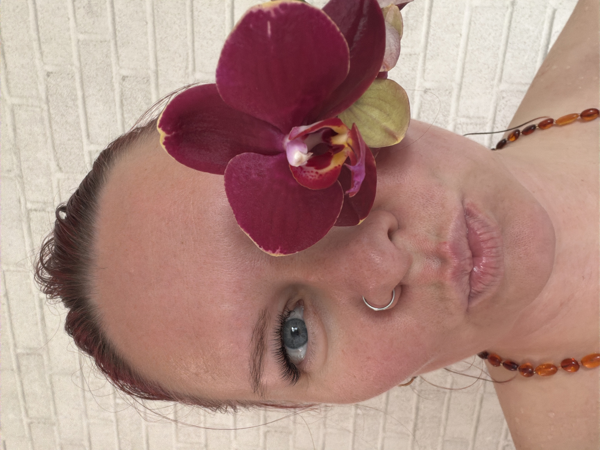 A close-up, slightly angled portrait features a person with fair skin and light blue eyes, adorned with a large, dark burgundy orchid placed above their left eye. A silver nose ring is visible. The person is wearing a necklace of amber-colored beads. The background appears to be a textured, off-white wall that resembles brick. The image does not provide sufficient information to determine that the location is in the city of Chaikovsky, Russia.
