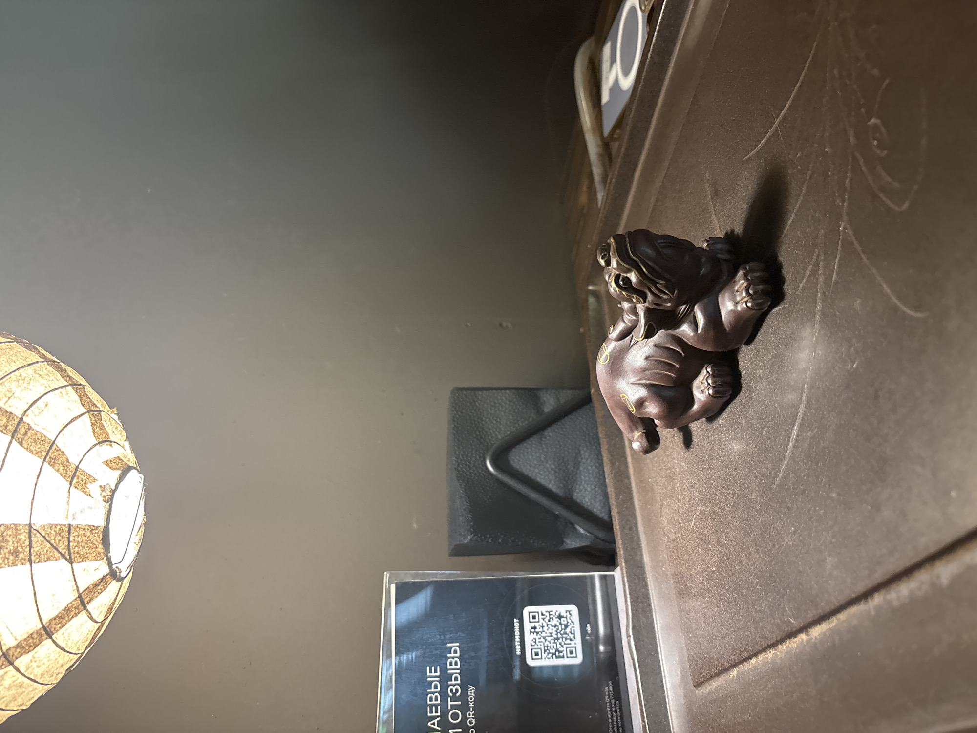 The image depicts an interior scene, possibly a cafe or restaurant, in Novaya Usman, Russia. In the foreground, a dark brown, carved figurine of a mythical creature, likely a dragon or lion-dog, rests on a textured dark brown tray. The figurine has gold-colored accents on its face and paws. To the left, a transparent display holder contains a QR code and text in Russian, which reads "МАЕВЫЕ ОТЗЫВЫ" and "С QR-коду". Adjacent to the display, a black napkin holder with black napkins is visible. Above the objects, a paper lantern light fixture casts a warm glow. The background is a plain, dark wall.