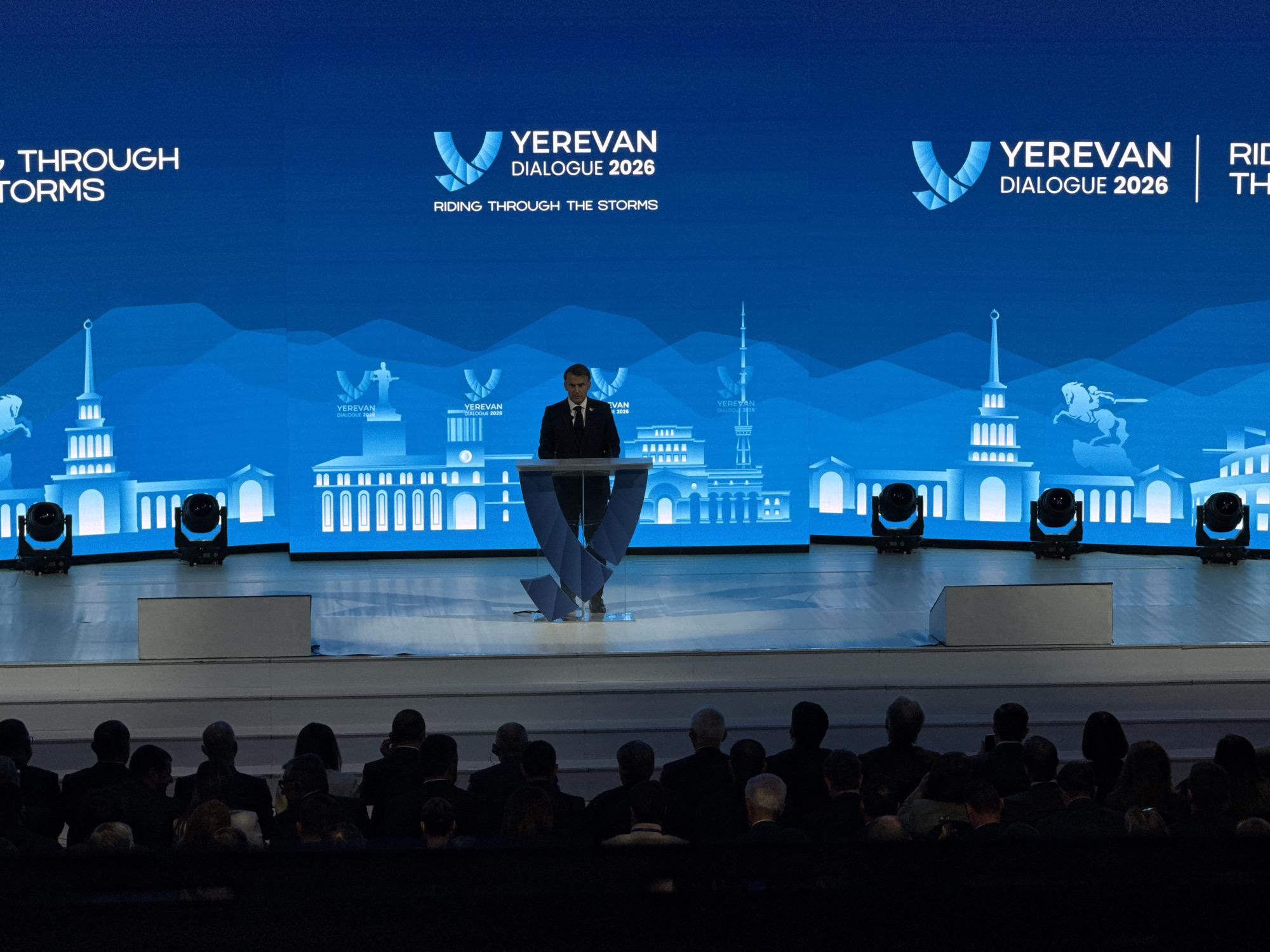 The image shows a man in a dark suit standing at a podium on a stage, addressing an audience. Behind him is a large blue screen displaying the words "Yerevan Dialogue 2026" and the phrase "Riding Through the Storms." The backdrop also features a stylized illustration of Yerevan's cityscape. Four stage lights are placed in front of the screen. The audience is seen in the foreground, their faces obscured in shadow.