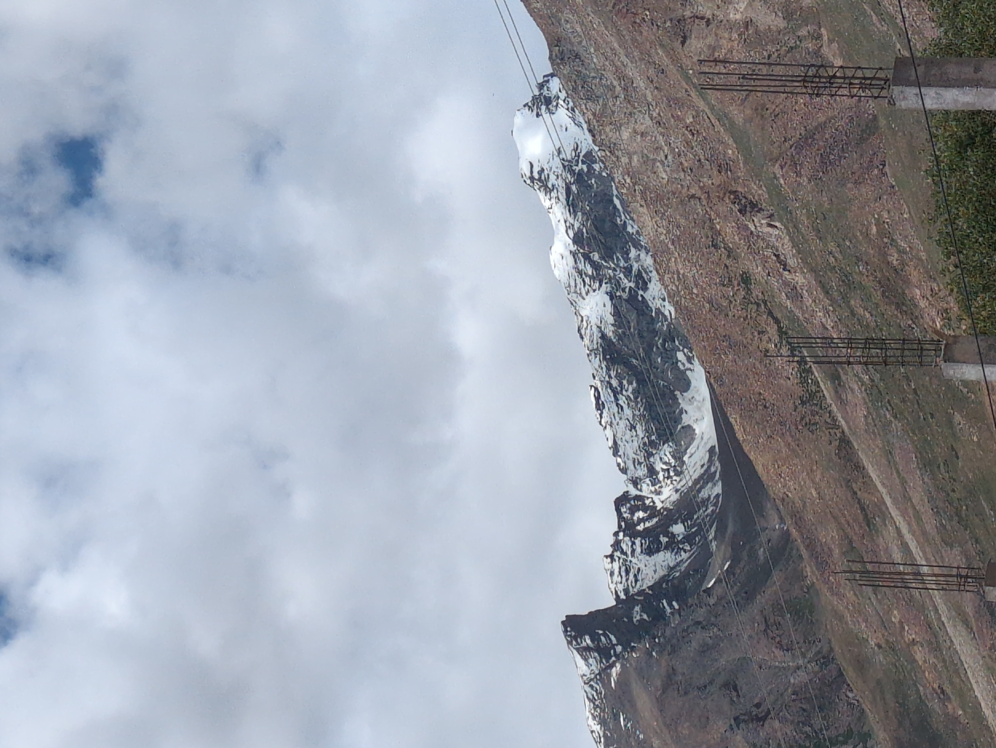 The image depicts a panoramic view of snow-capped mountains under a partly cloudy sky. The mountains are rugged and rocky, with patches of snow visible on their peaks and slopes. The foreground shows a steep, barren hillside with scattered vegetation and some construction materials, including rebar cages and concrete pillars, suggesting ongoing development or infrastructure work. Two power lines are also visible crossing the frame diagonally from right to left. The scene is likely situated in or near the city of Padum, India, a region known for its mountainous terrain and high altitude. The lighting suggests it is daytime, with the sun casting some shadows but not fully illuminating all parts of the landscape. There are no people visible in the image.