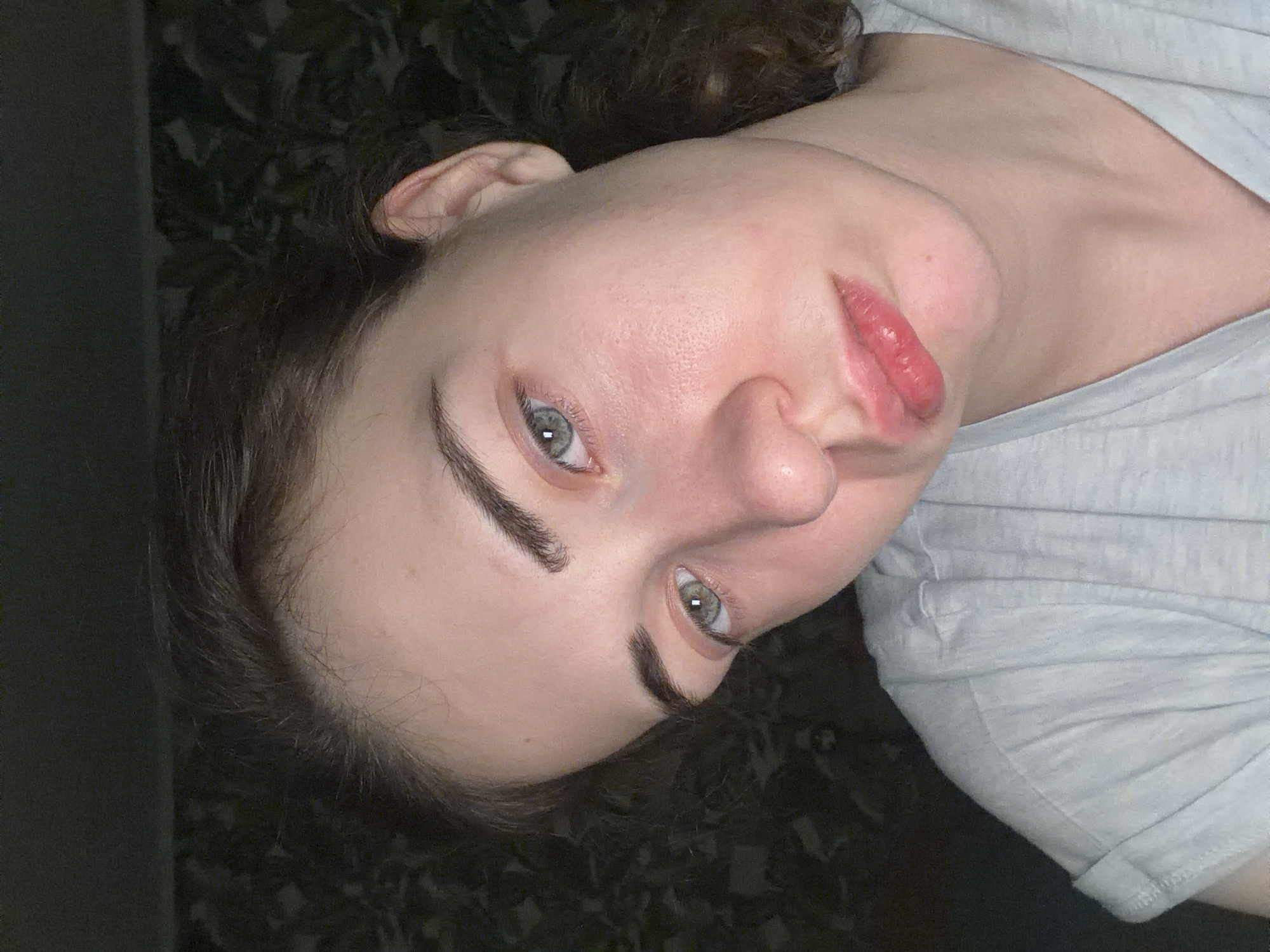 The image shows a close-up, head-and-shoulders portrait of a young woman with light skin and brown hair. She is wearing a light blue t-shirt. Her head is tilted, and she is looking slightly downward and to her left. Her eyes are green, and her lips are slightly parted. The background appears to be a dark, textured wall. There is no indication of the city of Zavodoukovsk, Russia, in this image.