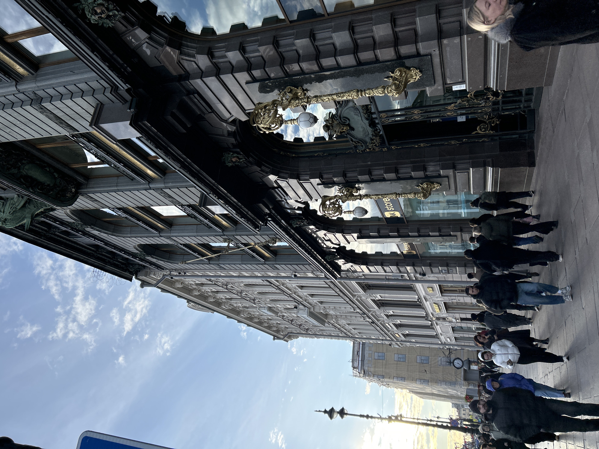 The image captures a vibrant street scene in Saint Petersburg, Russia, under a clear sky dotted with white clouds. The view looks down a wide city street lined with ornate architecture.
On the left side, a grand, dark-colored building with a richly decorated facade is prominent. It features dark grey stonework, intricate gold-colored decorative elements, and large, reflective windows. Ornate light fixtures with white globes extend from the building's exterior. Reflected in one of its windows, the inverted and mirrored text "BOOKSTORE" (or "K STORE") can be seen. Higher up, decorative carvings, possibly gargoyles or stylized faces, are visible.
Across the street to the right, a lighter-colored building, likely white or light grey, displays classical architectural details such as cornices, pilasters, and decorative window frames. Further down the street, another building appears, with a visible clock face on its facade and what seems to be scaffolding or construction elements on its roofline.
Numerous pedestrians are walking along the wide sidewalk, dressed in coats and jackets, suggesting cool weather. They appear engaged in their daily routines, with some figures blurred due to motion. In the immediate foreground on the far left, a portion of a blue road sign is visible. The sun, low in the sky, casts a bright glare and long shadows towards the right side of the street, indicating either late afternoon or early morning. The scene conveys the bustling atmosphere of a historic European city.