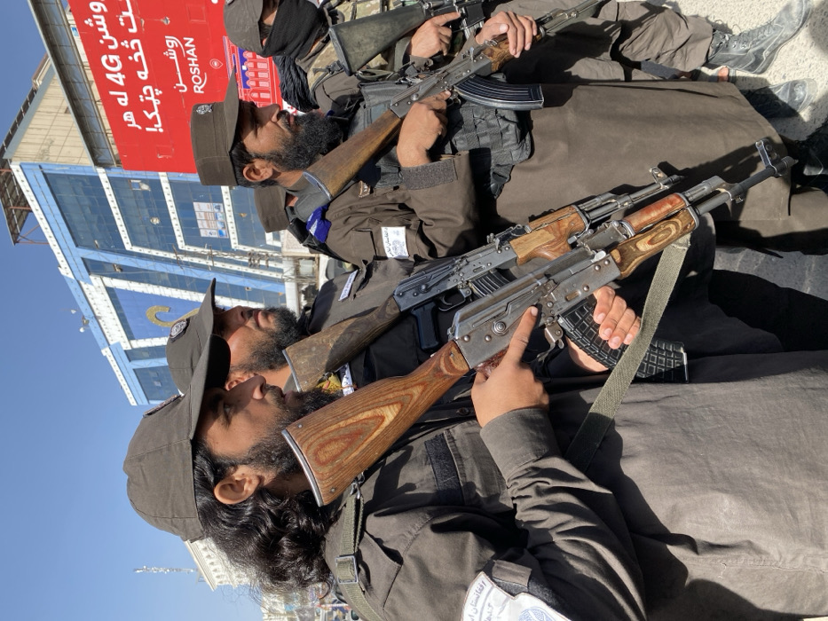 The image depicts a ground-level view of at least five armed men standing in an urban area of Kandahar, Afghanistan, on a clear, sunny day.
In the foreground, from left to right, are three prominent figures. The leftmost man has long, dark, curly hair, a full beard, and wears a dark olive-green baseball cap and a matching long-sleeved tunic. A white armband with blue script, consistent with local languages, is on his left upper arm. He holds an AK-style assault rifle with a wooden stock and handguard, and a black polymer magazine, with his right hand on the pistol grip and his left hand supporting the forestock.
Behind him, partially obscured, is another man with a dark beard and cap, also holding an AK-style rifle. To the right, a third man wears a dark brown garrison cap, a dark brown uniform (possibly shalwar kameez), and a black tactical vest or chest rig. A blue shoulder patch is visible on his right shoulder. He holds an AK-style rifle with a wooden stock.
Further right, two additional men are partially visible. One wears a dark uniform and tactical vest, holding an AK-style rifle. The man on the far right is mostly obscured, but wears a dark face covering, a dark cap, and holds an AK-style rifle. All visible men have beards and appear to be standing in a vigilant posture, indicative of public presence or patrol.
The background features a multi-story building with a facade of blue and white/silver rectangular panels. Above this, a large red billboard is visible, displaying white text in Dari or Pashto, including "4G" and the "ROSHAN" telecommunications company logo. The ground appears paved, suggesting a street or public thoroughfare.