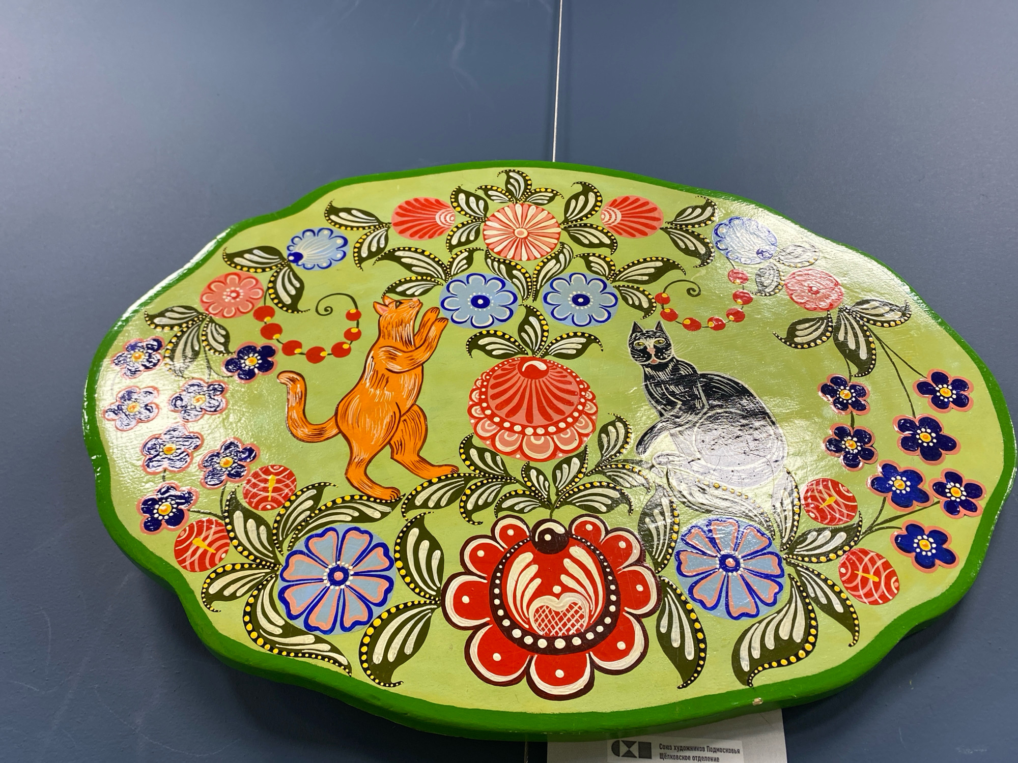 The image shows a close-up, eye-level view of a green, oval-shaped decorative plate, likely made of wood, displayed against a dark blue wall. The plate is adorned with a vibrant, folk-art style painting featuring two cats and floral motifs. An orange cat, standing on its hind legs with its front paws raised, is depicted on the left side of the plate. To the right of the orange cat is a black and white cat, sitting with its tail curled. The cats are positioned amidst an array of colorful flowers and leaves painted in red, blue, pink, and white with black outlines and yellow accents. The overall design has a whimsical, stylized appearance. Below the plate, a portion of a white label is visible, with text that appears to be in Cyrillic script, suggesting the plate is an exhibit. Based on research, this artwork is likely located at the Ivanteyevka Museum of Local Lore, which is situated in the city of Ivanteyevka, Moscow Oblast, Russia.