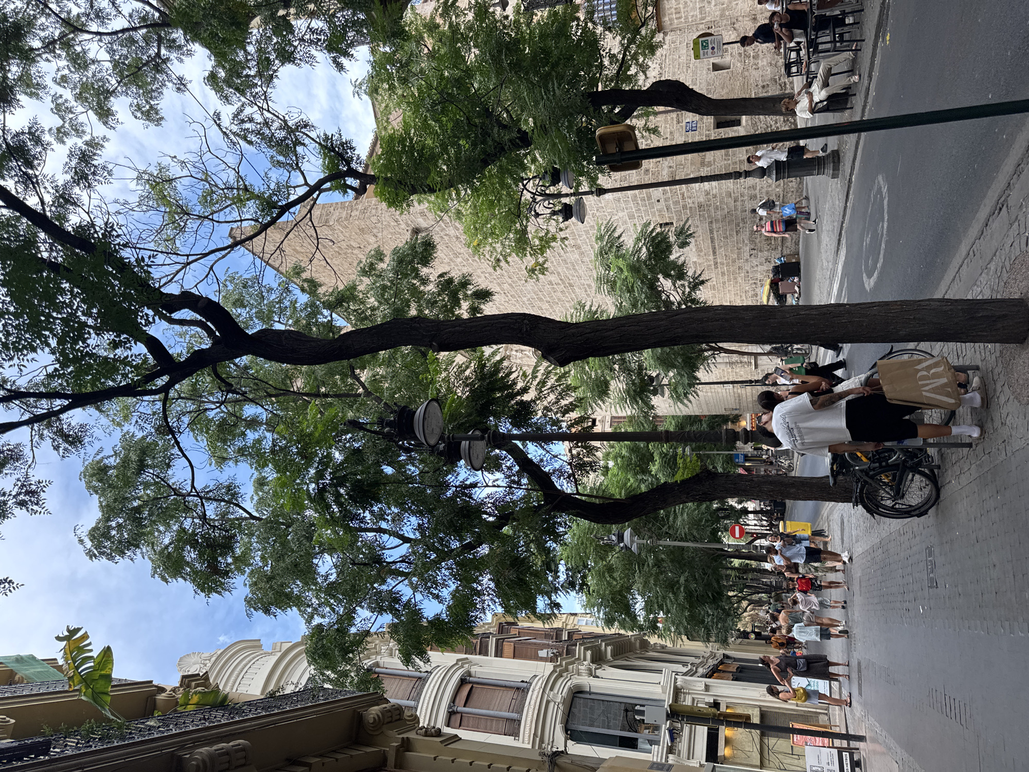 This image depicts a street scene in the city of Valencia, Spain, likely within a historic district given the architecture. The foreground features a paved sidewalk lined with mature trees casting shadows on the ground. Several individuals are walking along the sidewalk. On the left, a white building with ornate architectural details and curved windows is visible. To the right, a brick building with arched windows forms the backdrop.
In the middle ground, a group of people, including children, are walking away from the camera. A bicycle is parked on the sidewalk near a lamppost. A shopping bag with the "ZARA" logo is held by a person in the foreground. Further down the street, more people are visible, some sitting at outdoor tables of a cafe. Traffic lights and street signs are also present. The sky is partially cloudy. The overall impression is that of a bustling urban environment on a clear day.