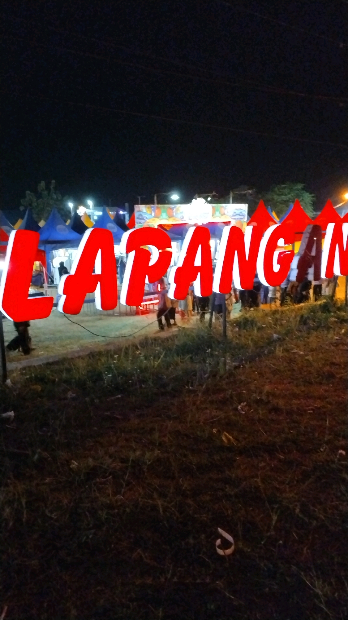 At night, a large red sign with white outlining is illuminated, spelling out "LAPANG AN" in bold, three-dimensional letters. The sign stands prominently in the foreground, with a grassy area in front of it. In the background, several tents, likely part of a market or event, are visible under the night sky, some with colorful banners. Lights from the tents and streetlights illuminate the scene. People are seen walking around the tents and in front of the sign, suggesting an outdoor gathering or festival. The overall impression is of a lively night market or fairground. The image captures an outdoor scene at night, possibly in Canggu, Indonesia, featuring a large illuminated sign for a "LAPANG AN" (likely meaning "field" or "area" in Indonesian) event or establishment. Several tents are set up in the background, indicating a market or festival atmosphere. Some individuals are present, milling about the area.