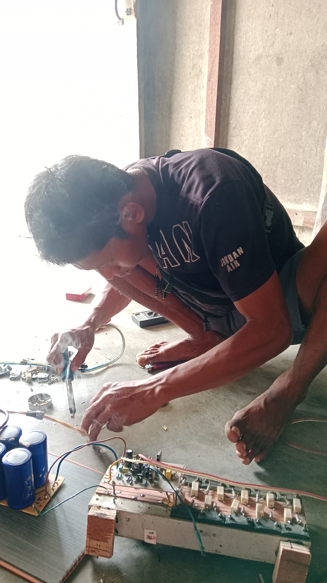 In Karang Indah, Indonesia, a person is crouched on the floor, engaged in soldering electronic components. The individual wears a dark t-shirt and shorts, with a backpack strapped on. In front of them lies a circuit board populated with various components, including capacitors, resistors, and transistors, connected by wires. A soldering iron is being used, producing smoke, and a lighter is visible nearby. The scene appears to be an indoor workspace with a concrete floor and wall.