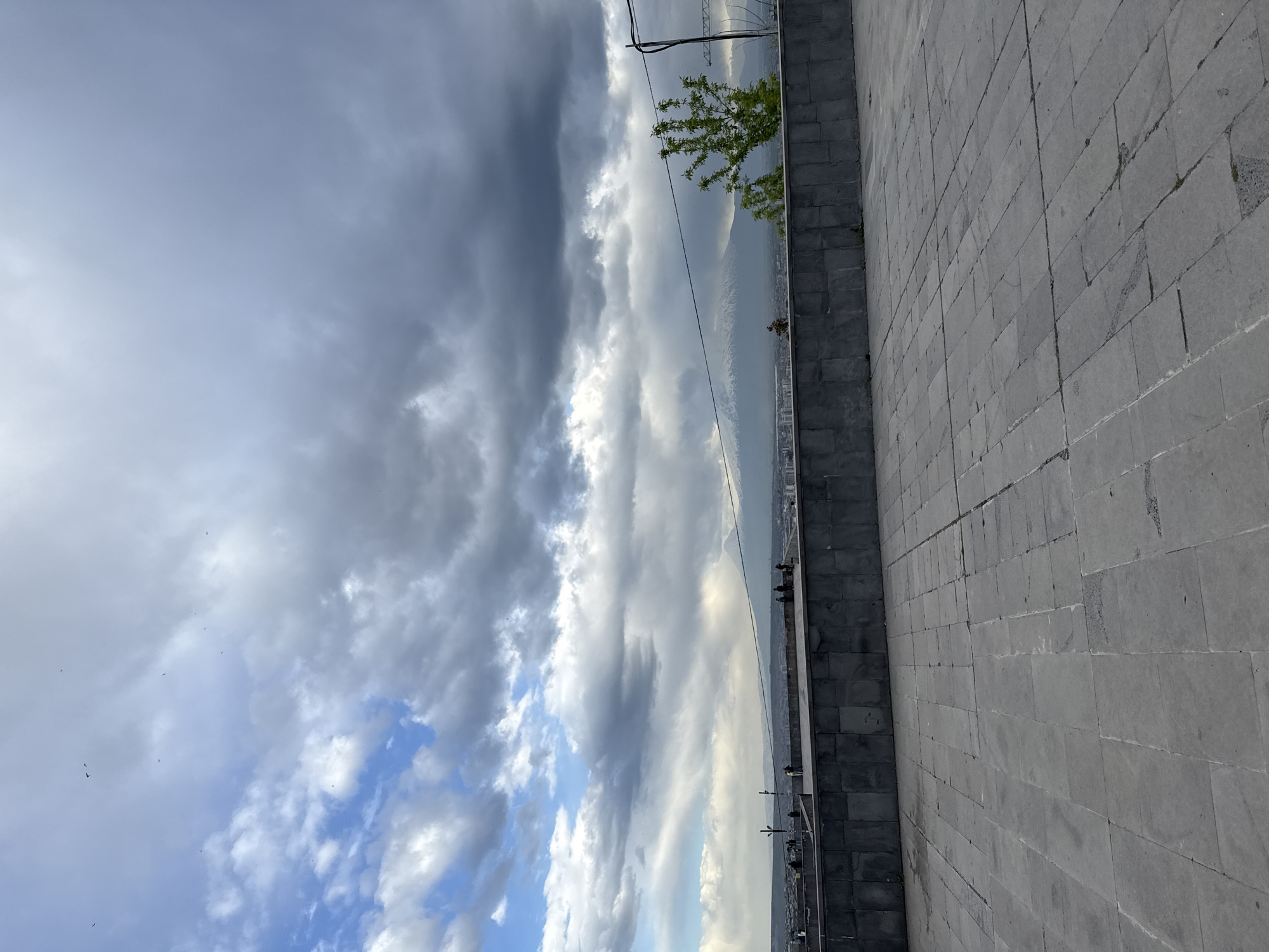 The image presents a view from a high vantage point in Yerevan, Armenia, likely from the Cascade complex. The foreground features a grey, stone-paved surface, possibly a walkway, transitioning into a raised grey stone retaining wall. Beyond the wall, a panoramic view unfolds, showcasing a city skyline partially obscured by a layer of haze or smog. Overlapping the cityscape is a backdrop of layered clouds in varying shades of grey and white, with patches of blue sky visible. A tree is positioned near the retaining wall with the foliage visible in the image. Powerlines can also be seen cutting through the scene. Several birds are also visible in the sky.