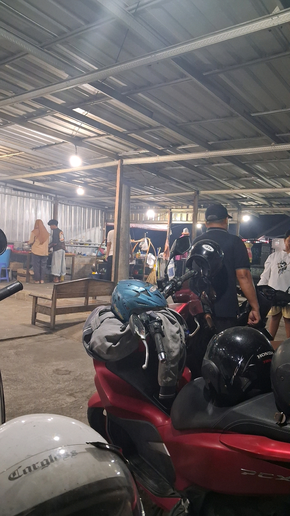 The image depicts an outdoor food stall or dining area in Berbah, Indonesia. The scene is illuminated by overhead lights, and a corrugated metal roof supported by wooden beams covers the area. In the foreground, two motorcycles are parked, one red and one white, each with helmets placed on the seats or handlebars. One helmet is blue, and another is black with the "HONDA" logo visible. In the mid-ground, several people are present. Two individuals, one wearing a hijab and a tan top, and another in a dark jacket and light pants, appear to be working or preparing food behind a counter. Another person in a white shirt stands nearby. Further in the background, more people are visible, and a variety of items, including what appears to be food stalls and cooking equipment, are set up. The overall atmosphere suggests a bustling, casual dining environment at night.