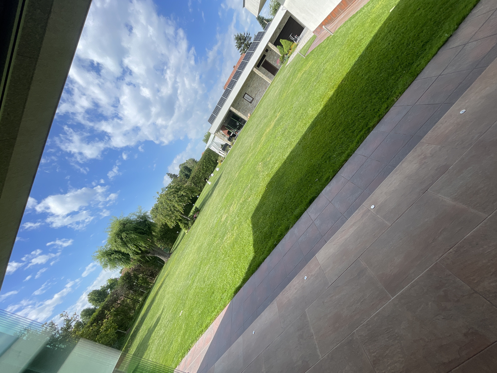 The image depicts an outdoor scene with a house, lawn, and patio. The lawn is green and well-maintained, with trees bordering the left side of the frame. A house is visible in the background with solar panels installed on the roof. To the right of the lawn is a tiled patio area. The sky is blue with scattered clouds. The image appears to be taken from indoors, looking out through a window or doorway. The location is Ryahovo, Bulgaria.