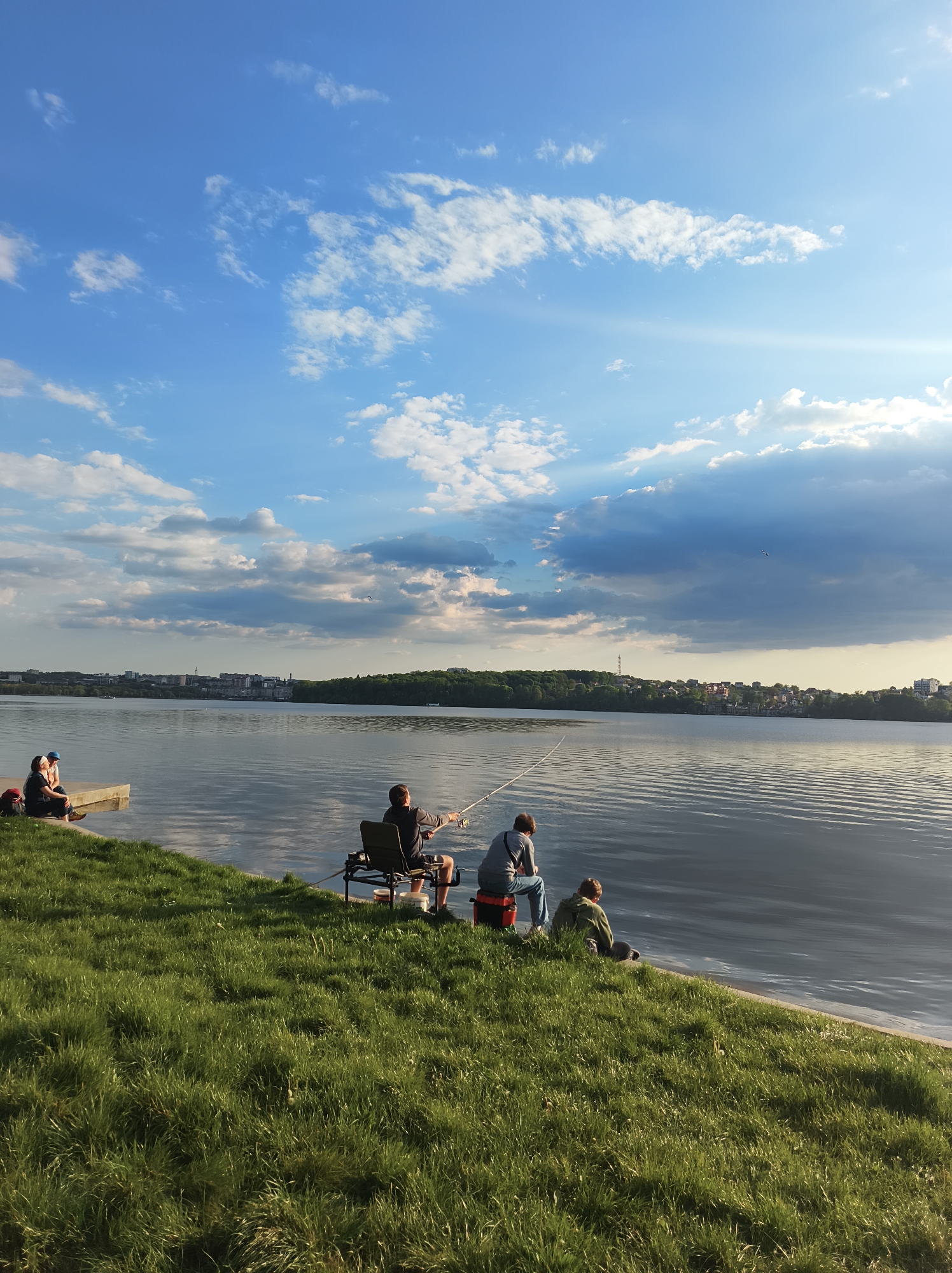The image depicts a lakeside scene in Ternopil, Ukraine. In the foreground, a grassy bank slopes down to the water's edge. A group of individuals is engaged in fishing. One man is seated in a chair with a fishing rod in hand, another person sits on a red box, and a third individual sits on the grass beside them. Further to the left, other people are seated on a concrete structure near the water. The water surface is relatively calm with visible reflections of the sky. In the background, a skyline of buildings and trees is visible along the horizon. The sky is partly cloudy with patches of blue, and sunlight is streaming through the clouds.