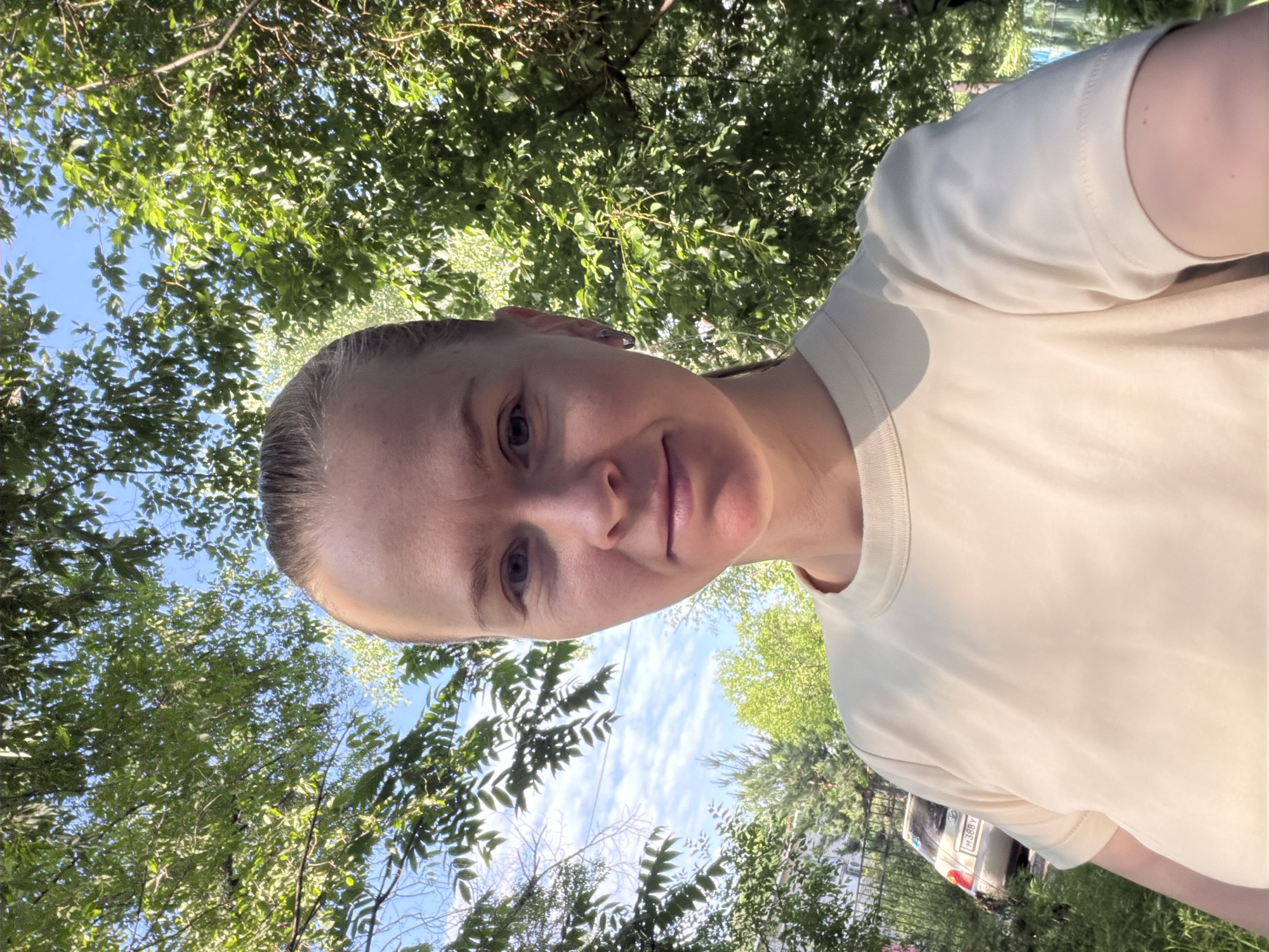 A young woman is taking a selfie outdoors. She has light skin, light hair pulled back in a ponytail, and is wearing a light yellow t-shirt. She has a small silver hoop earring in her left ear. She is positioned in the foreground, with green trees and blue sky filling the background. A parked car and a fence are visible behind her to the left. The lighting suggests it is daytime. This scene appears to be in an urban or suburban residential area, possibly in Khabarovsk, Russia, given the presence of a car with a Russian license plate.