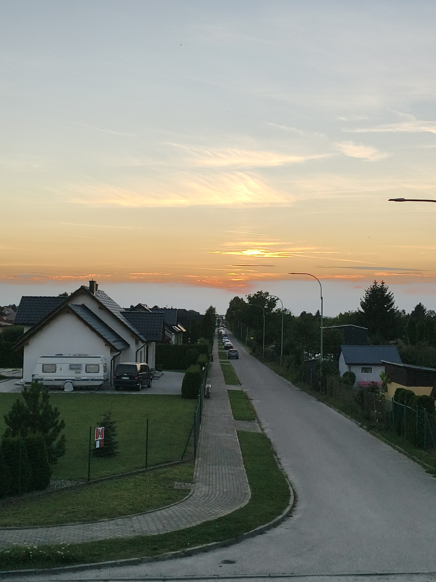 The image depicts a residential street in Tczew, Poland, during the golden hour of sunset. The sky is filled with soft, wispy clouds tinged with orange and yellow hues, indicating the sun is low on the horizon. The street is paved and curves gently to the right, lined with houses and trees. On the left side of the street, a white house with a dark tiled roof is visible, with a caravan parked in its driveway. A black SUV is also parked on the driveway. Adjacent to the house is a well-maintained lawn with a small evergreen tree and a fence. Further down the street, several cars are parked along the side. On the right side of the street, more houses and trees can be seen, including a smaller building with a dark roof. Streetlights are positioned along the road. The overall impression is one of a peaceful suburban evening.