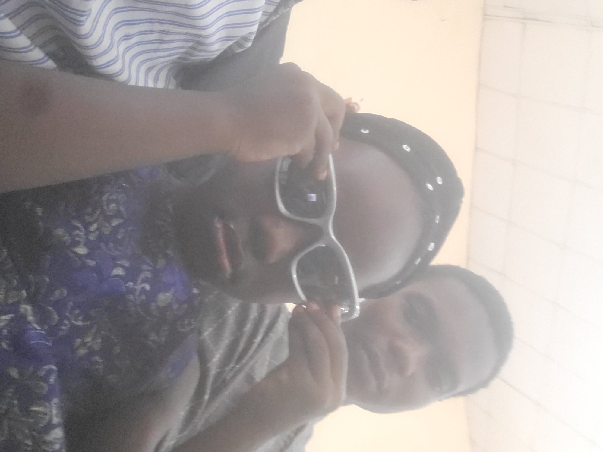 Two dark-skinned individuals are in the frame. One wears sunglasses and a beaded headscarf, partially obscured by their hand adjusting the sunglasses. They wear a dark, floral-patterned top. The second individual's face is partially visible behind them. They wear a grey shirt. A person wearing a striped shirt is partially visible at the top of the frame. The location appears to be indoors, with a light-colored wall and possibly tiled surfaces in the background. The city and specific location are unknown.