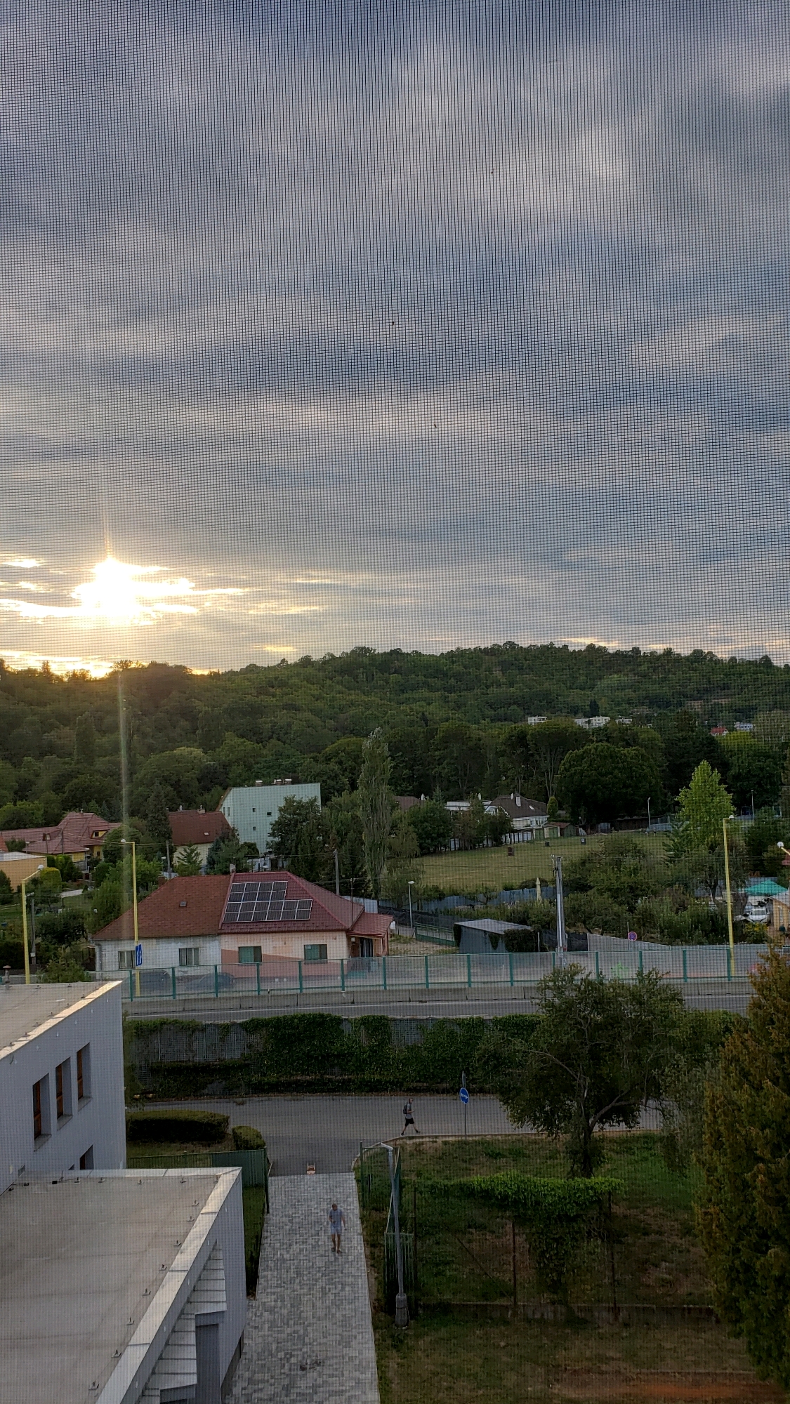 The image depicts an outdoor scene with a clear view of a residential area and a wooded hill. The sun is visible in the upper left quadrant, partially obscured by clouds, casting a bright light. Several houses with red roofs are scattered throughout the landscape, some featuring solar panels. A modern building with a flat, gray roof and multiple windows is in the foreground on the left. A paved walkway leads down from this building. Two individuals are present: one person is walking down the paved walkway, and another individual is jogging on a road in the middle ground. The environment includes lush green trees and vegetation, as well as a fence line and a road. The overall impression is that of a suburban or semi-rural setting. This scene appears to be located in the city of Prešov, Slovakia, possibly in a residential area near a hillside.