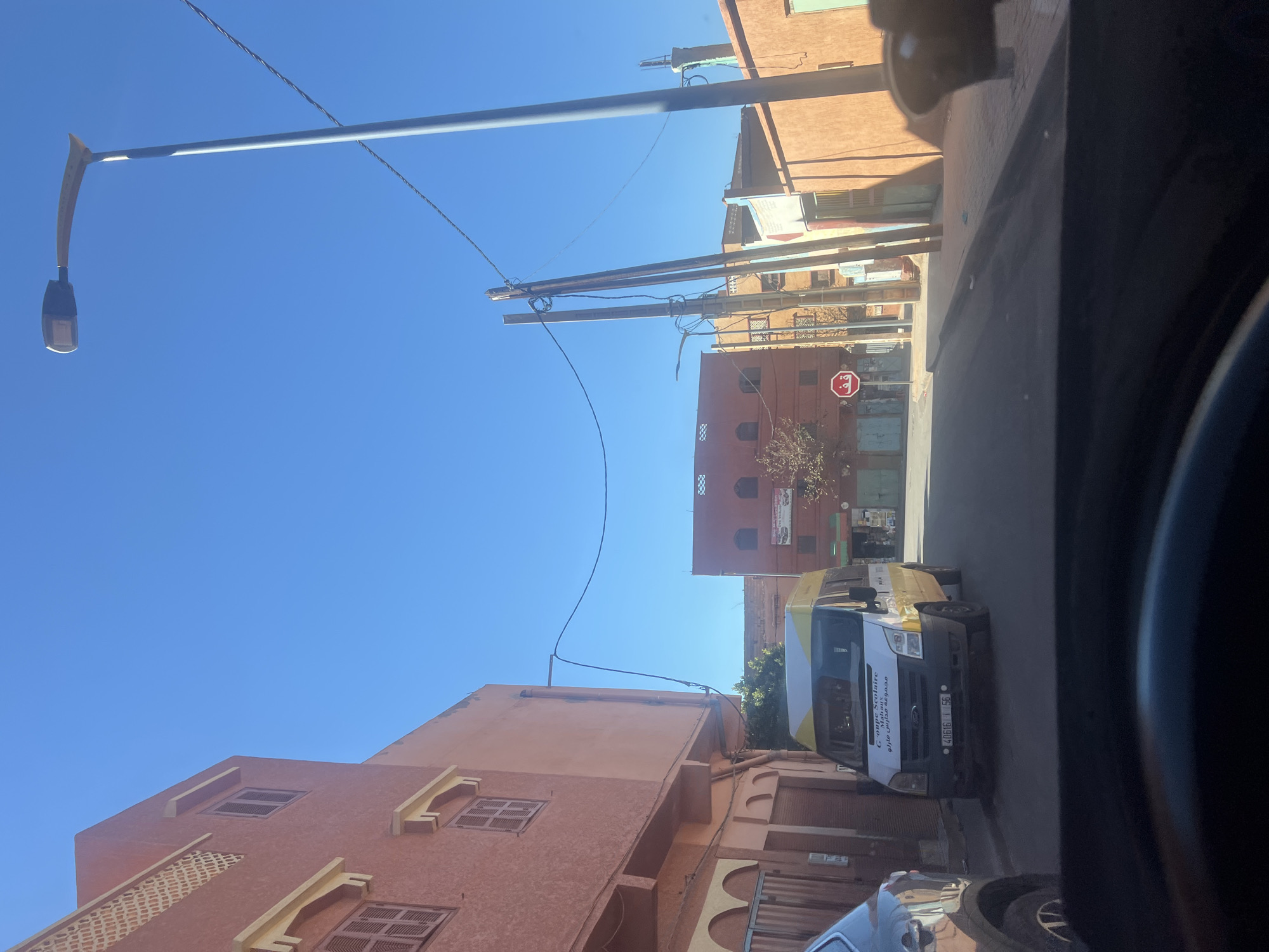 An eye-level shot from inside a vehicle in the Moroccan town of Ouaouizert reveals a typical street scene. The town's architecture is characterized by terracotta-hued buildings with a mix of window styles. A school bus is positioned on the street with the words "Groupe Scolaire" on its side. Above the buildings are electrical cables, street lamps, and a blue sky. A red stop sign is also visible on the side of a building.