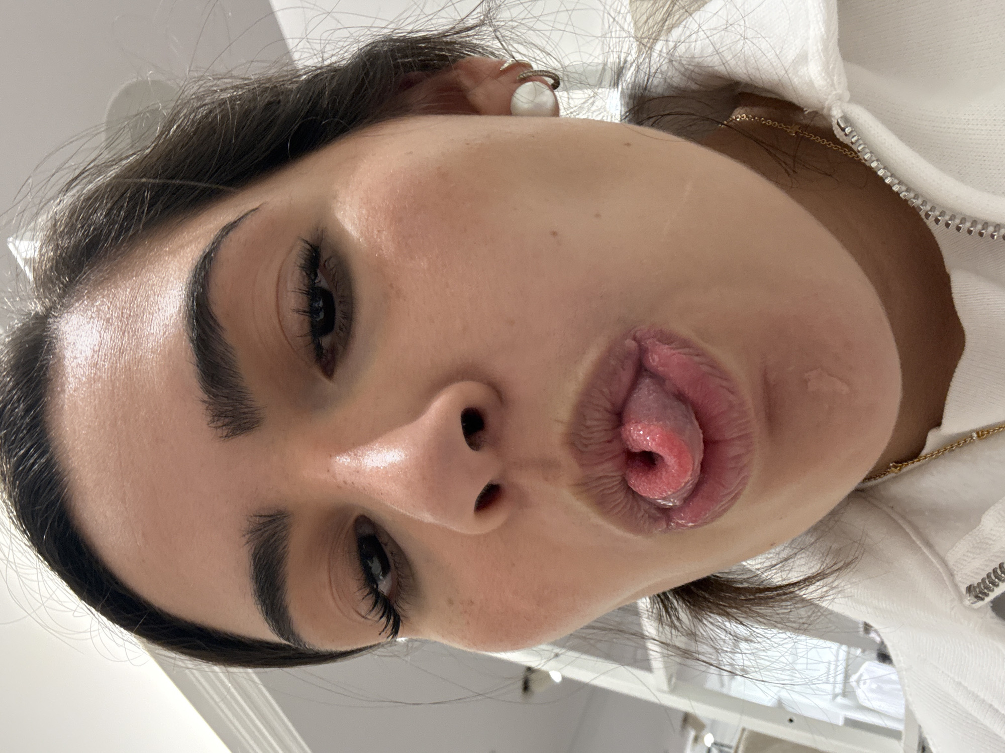A close-up, low-angle shot depicts a light-skinned individual with dark hair and prominent eyebrows. The person is sticking out their tongue and puckering their lips. They are wearing a white hooded garment with a gold chain visible at the neckline. An earring is visible on their left ear. The background is blurry and doesn't provide any contextual information to discern the specific location in San Antonio, USA. The focus is on the person's facial expression and features.