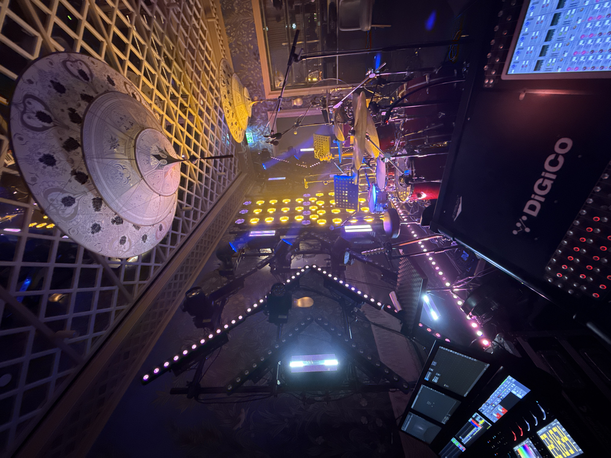 The image depicts a dimly lit indoor music venue, likely a club or small concert hall, in Baku, Azerbaijan. The foreground features a professional audio mixing console, branded "DiGiCo," with multiple screens displaying waveforms and faders, indicating it is set up for a live performance. Behind the console, a drum kit is visible, complete with cymbals, drums, and microphones, suggesting a musical act is either present or about to perform. The background is adorned with decorative ceiling elements and wallpaper, hinting at a sophisticated or themed interior. Stage lighting, including LED strips and spotlights, illuminates the area with vibrant purple and yellow hues, creating a dynamic atmosphere. The overall scene is a backstage or performance setup, ready for a musical event.