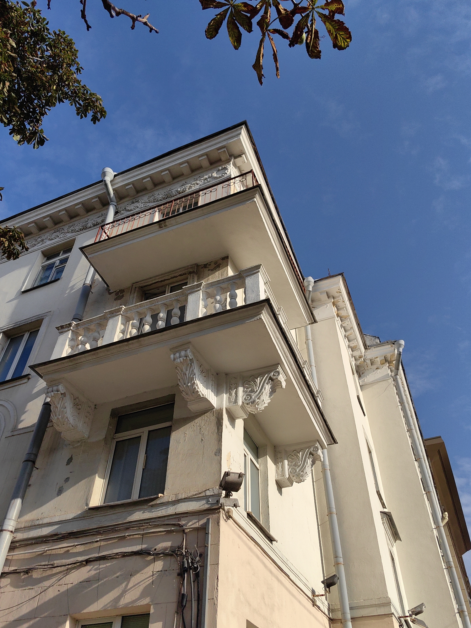 An upward-facing, wide-angle, full shot captures a light-colored building with balconies against a bright blue sky with wispy clouds. Branches of a horse chestnut tree with green and yellowing leaves are visible in the upper left and upper right corners. The building features decorative white plasterwork on the balconies, cornices, and along the roofline. The balconies have white balustrades with turned spindles and are supported by ornate brackets. Downspouts run vertically along the building's facade. Windows with white frames are visible on multiple levels. The building is situated in the city of Minsk, Belarus.