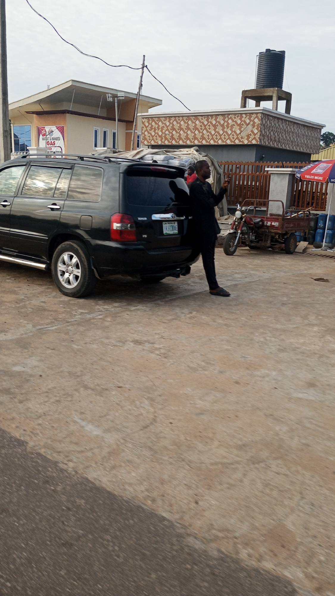 The image presents an outdoor scene in Lokoja, Nigeria, under an overcast or hazy daytime sky. The ground is a mix of light concrete and a darker, possibly asphalted, surface.
In the mid-ground, a black Toyota Highlander SUV is prominently parked, showing its rear. The license plate displays "LAK 350MC," and "TOYOTA," "HIGHLANDER," and "V6" badges are visible. Bags, possibly containing goods, are piled on its roof rack. A man, dressed in dark clothing, is standing next to the rear passenger side of the SUV, seemingly interacting with a mobile phone.
In the background, two buildings are visible. On the left, there's a light-colored building with a modern roofline and blue-tinted windows. A prominent white sign with red text on this building reads "HAFSAT & HAMEED GAS." To the right, another building features an ornate facade of brown and beige patterned tiles, topped with a black water tank on a concrete support structure.
Further right in the mid-ground, a red motorcycle with a three-wheeled cargo trailer is parked. Nearby, several blue barrels are stacked, and a small red and blue umbrella bears the text "YIELDS MORE." Power lines are visible stretching across the upper portion of the frame. The scene suggests a busy commercial or residential area with various ongoing activities.