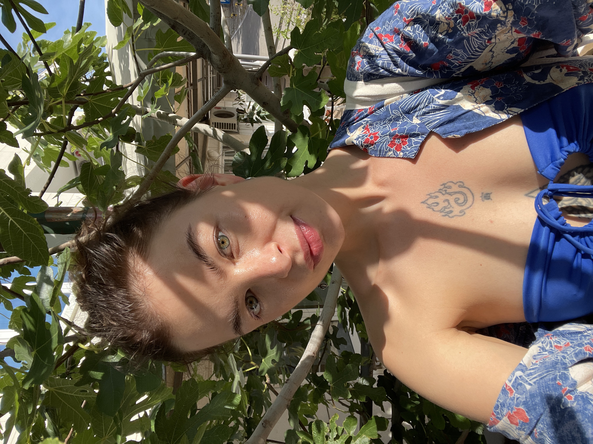 A close-up, angled, and brightly lit photo captures a person posing outdoors, surrounded by lush green foliage. The person, positioned upside down in the frame, looks directly at the camera with a slight smile. Their face is partially shaded by leaves and branches, creating dappled sunlight patterns. They have striking green eyes and a natural, light makeup look with glossy lips. Dark, short hair frames their face.
They are wearing a blue swimsuit top, partially visible, and a loosely draped, open blue and white patterned garment with red accents over it. A small, stylized tattoo of a cloud and some text are visible on their chest. The background consists of green fig leaves and branches, with glimpses of a building structure and what appears to be a clear sky. The bright sunlight suggests it is daytime, possibly morning or afternoon. The overall impression is one of a relaxed, natural setting.