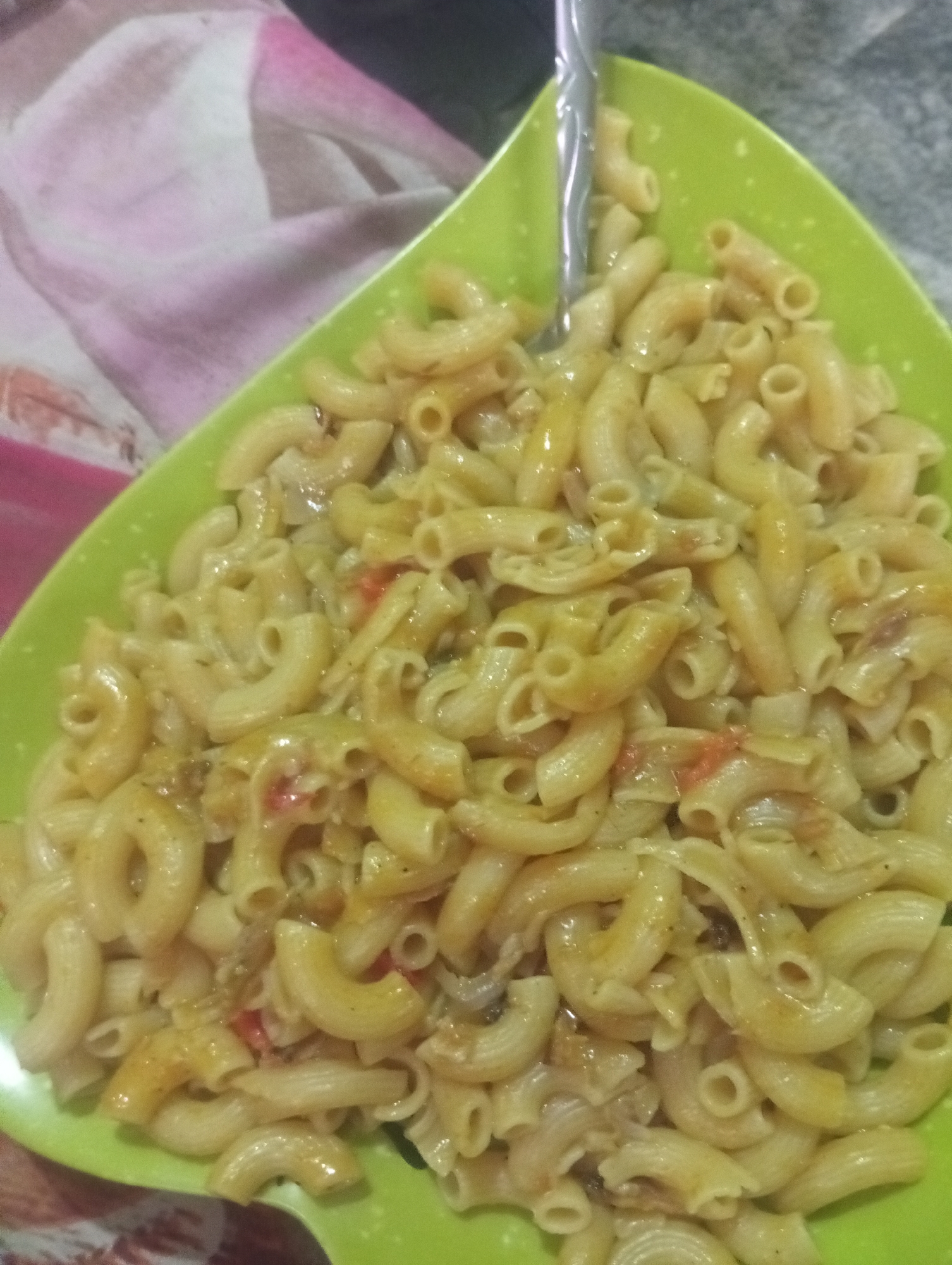 A close-up shot shows a green, triangular plate filled with macaroni pasta. The macaroni appears to be coated in a light sauce, with pieces of what might be tomatoes and onions visible amongst the pasta. A metal fork is inserted into the pasta. The plate sits on a surface covered with a patterned cloth of pink, white, and red hues. The image quality is somewhat grainy. Information related to location cannot be confirmed based on the image.