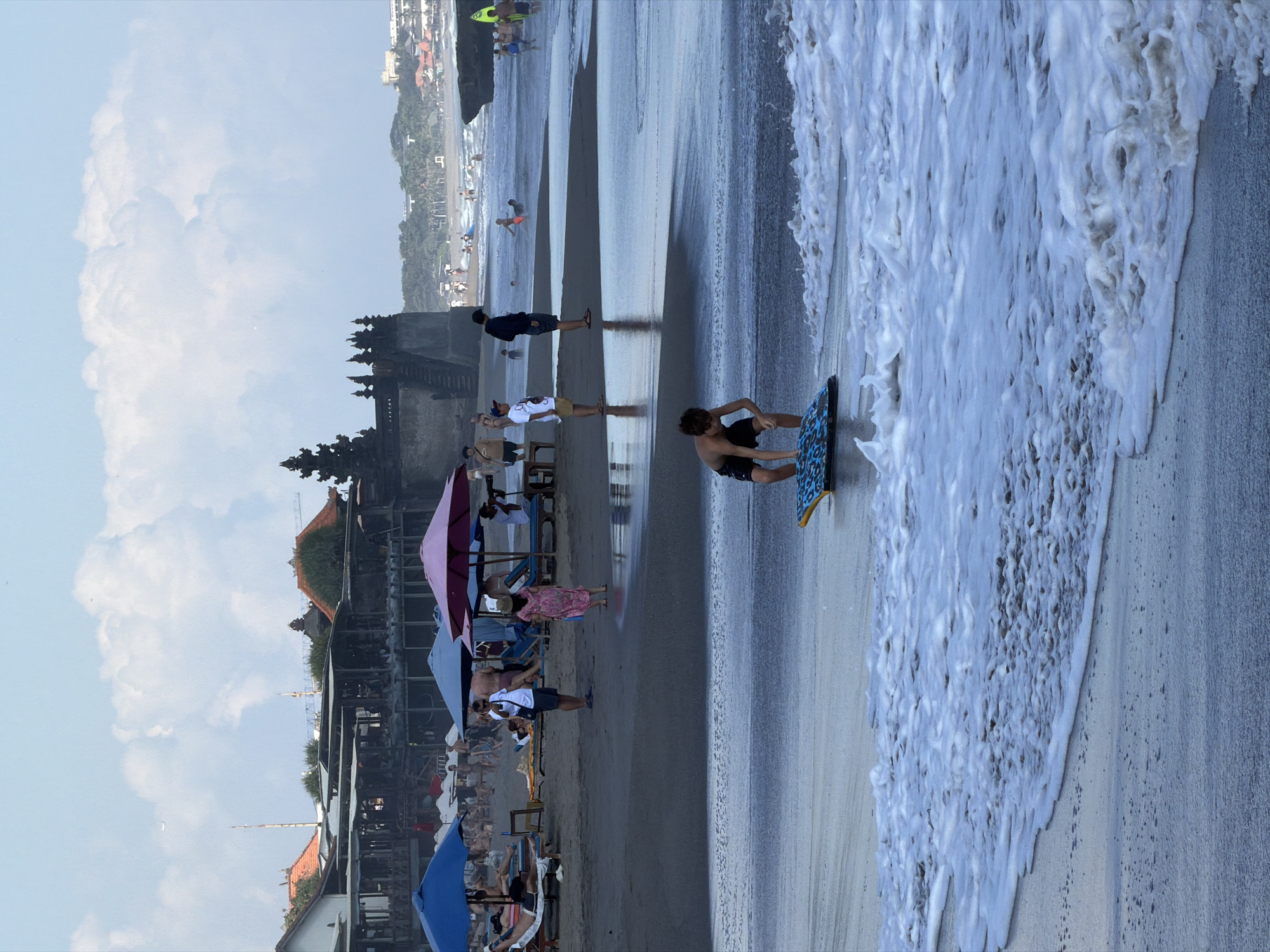 The image depicts a beach scene in North Kuta, Indonesia, likely during the daytime with overcast skies. In the foreground, a young boy is crouched on a boogie board in shallow water with frothy waves. The wet sand reflects the sky. Further back on the beach, several people are visible, some lying on lounge chairs under umbrellas, others standing or walking. Beach umbrellas in blue, pink, and purple are scattered across the sand. In the mid-ground, there is a line of buildings that appear to be restaurants or shops, with thatched roofs and open-air seating areas. A distinctive tiered temple structure with dark stone is situated on higher ground behind the buildings. In the distance, more people are seen in the ocean and on the shoreline, with a rocky outcrop to the right and a hazy coastline beyond. The overall impression is of a popular tourist beach with a mix of relaxation and water activities.