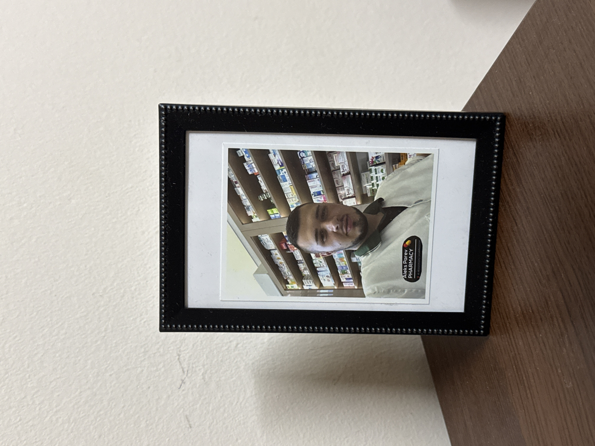 A framed photo hangs on a wall with two distinct surfaces. The photo features a man in what appears to be a pharmacy setting. Shelves stocked with various packaged medications are visible behind him. The man is wearing a white shirt with a black collar and a logo that says "Aleks Parev Pharmacy". The frame is black with a beaded edge. The wall is divided into two textures, a textured white surface on the left and a smooth brown surface on the right. Based on the logo, this photograph may have been taken in Sofia, Bulgaria, where Aleks Parev Pharmacy is located.