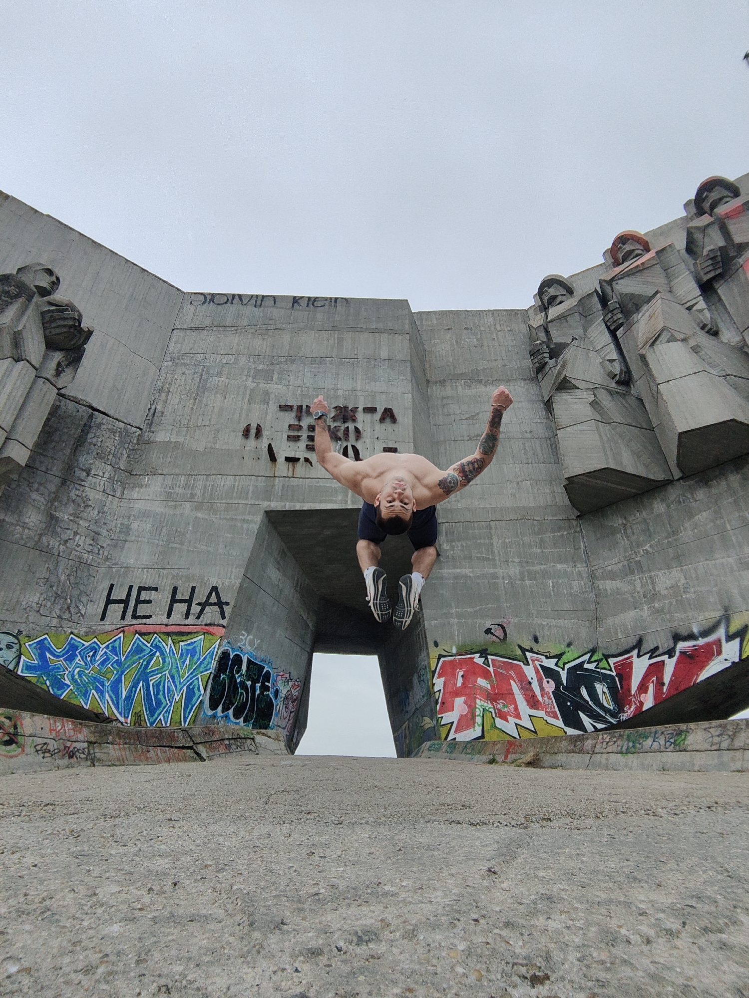 A person is captured mid-air, performing a backflip at the Monument of the Bulgarian-Soviet Friendship in Varna, Bulgaria. The monument, composed of grey concrete, features a central arched opening and is adorned with sculptural relief figures. Graffiti is visible on the lower portions of the monument. The surface in the foreground appears to be concrete or stone. The sky above is overcast.