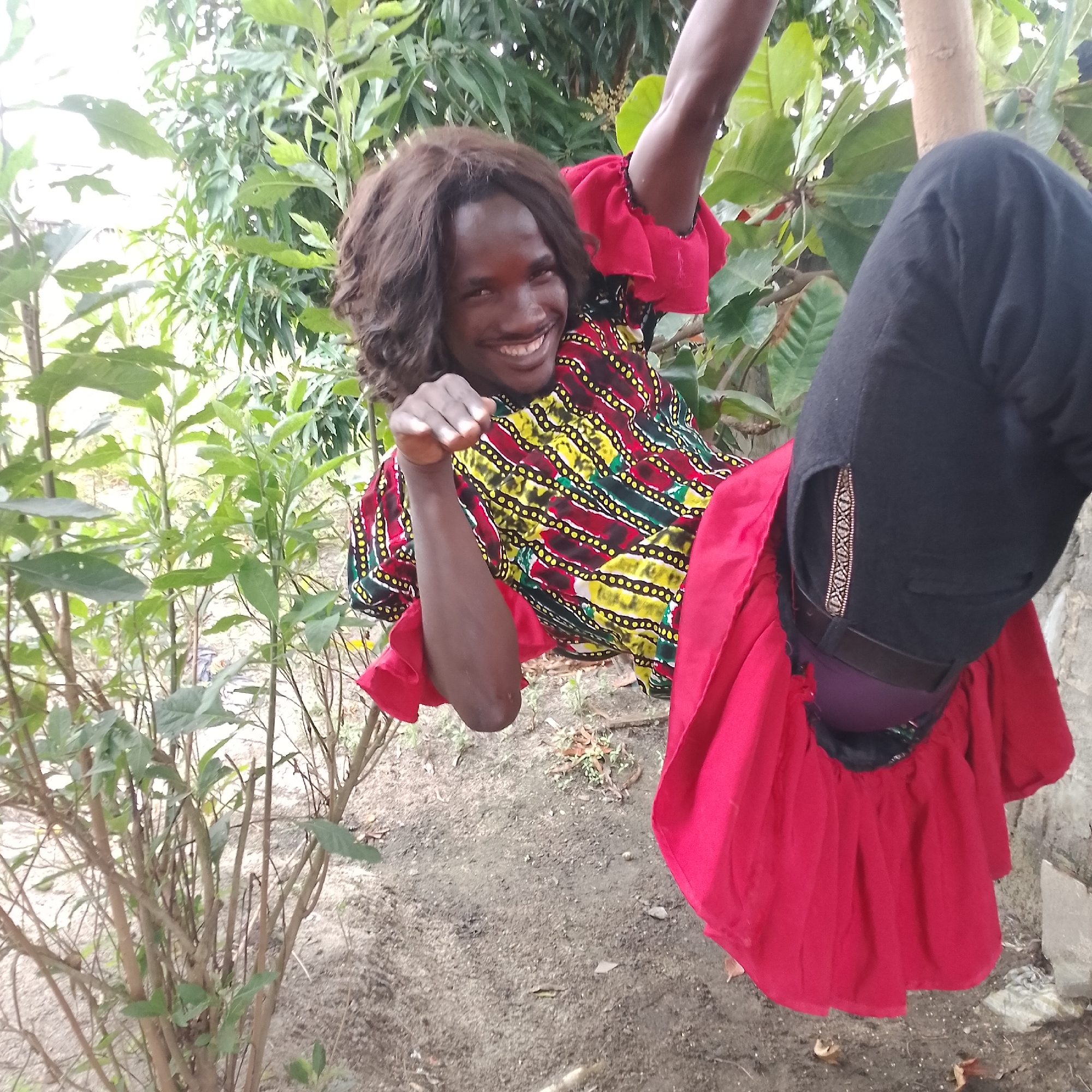 An individual with dark, curly hair and a wide smile is partially visible in the upper portion of the frame, in Ado, Nigeria. The person is wearing a colorful, patterned garment with red ruffled sleeves and black trousers. Their right arm is raised, and their left arm is bent with their hand near their face. The subject appears to be leaning against a wooden pole, with their legs bent and a large red fabric draped around them. The background consists of lush green foliage and trees, with some dirt and debris on the ground. The lighting suggests it is daytime.