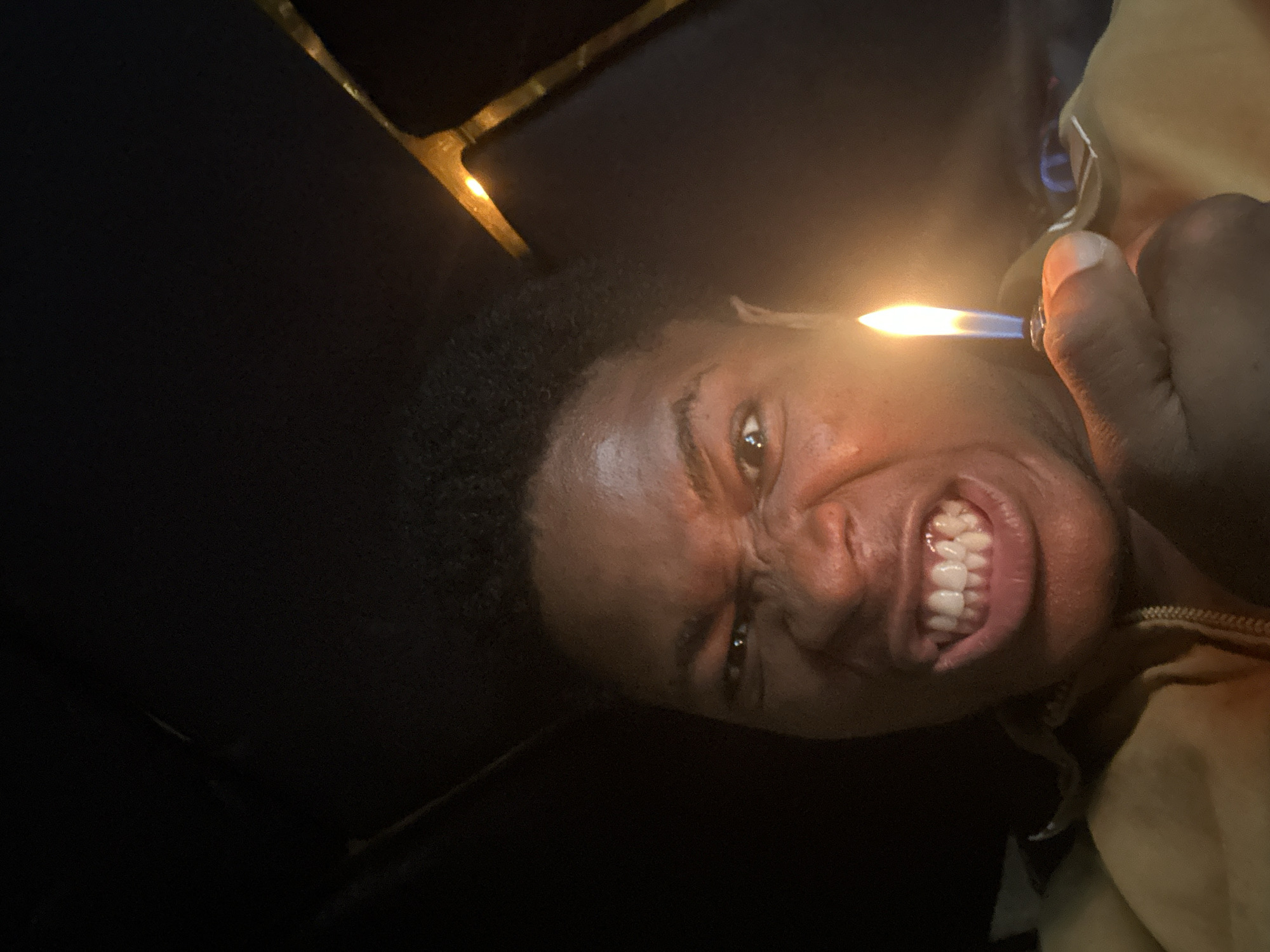 A close-up selfie shows a Black individual with a grimacing expression, holding a lit lighter in their right hand. The flame of the lighter illuminates the lower half of their face, highlighting their teeth and lips. The background is dark, with some indistinct yellow or orange shapes visible on the right side, possibly indicating reflections or an object in the environment. The individual is wearing a light brown jacket. The image appears to have been taken in Ihiagwa, Nigeria, though no specific landmarks or contextual details of the location are discernible within the frame.