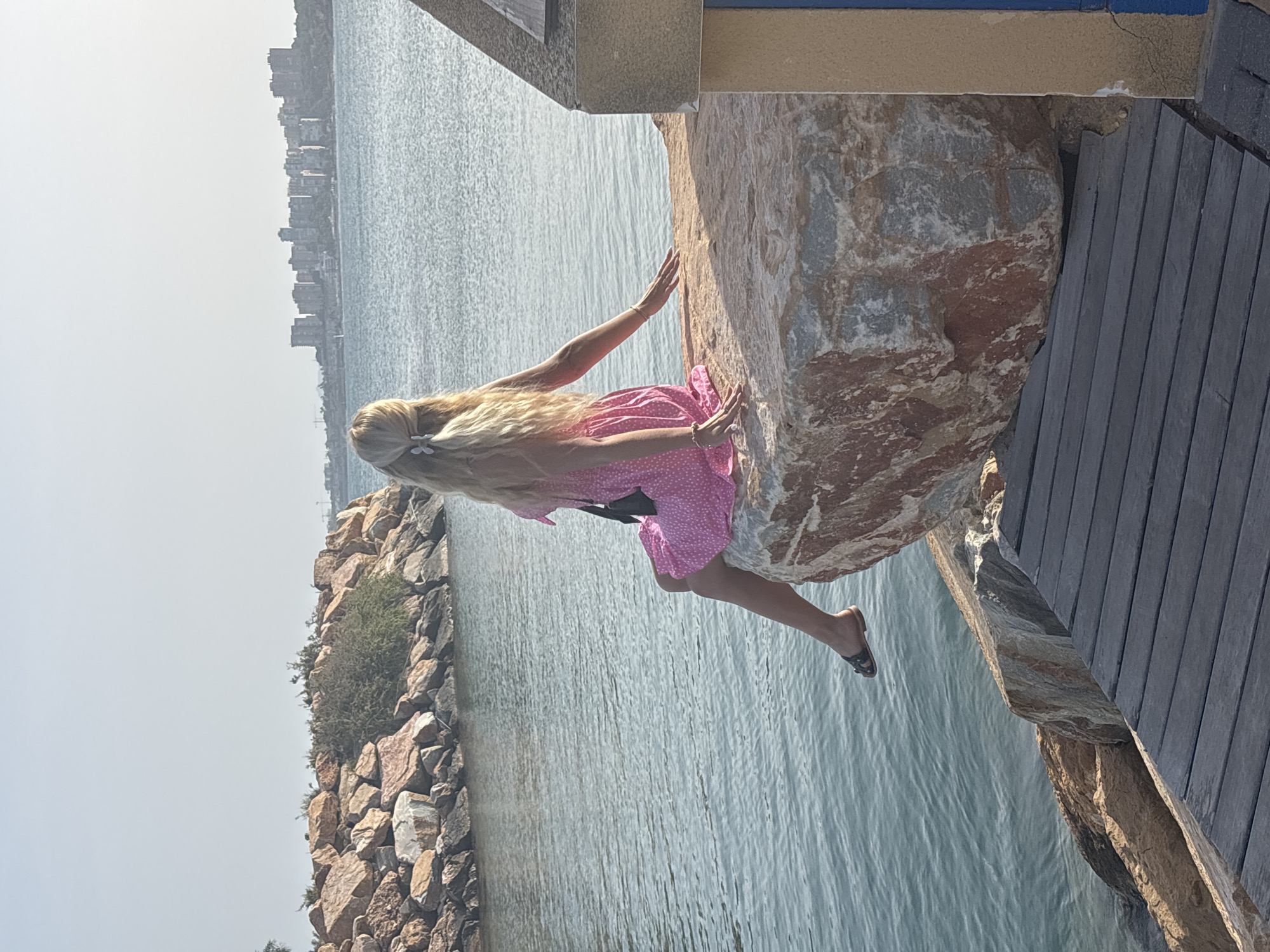A woman with long blonde hair is sitting on a large rock by the water. She is wearing a pink polka dot dress and black sandals. Her hands are resting on the rock. The scene appears to be a waterfront with a city skyline in the background. The water is calm and the sky is bright, suggesting a sunny day. The woman's pose and the setting evoke a sense of leisure and relaxation.