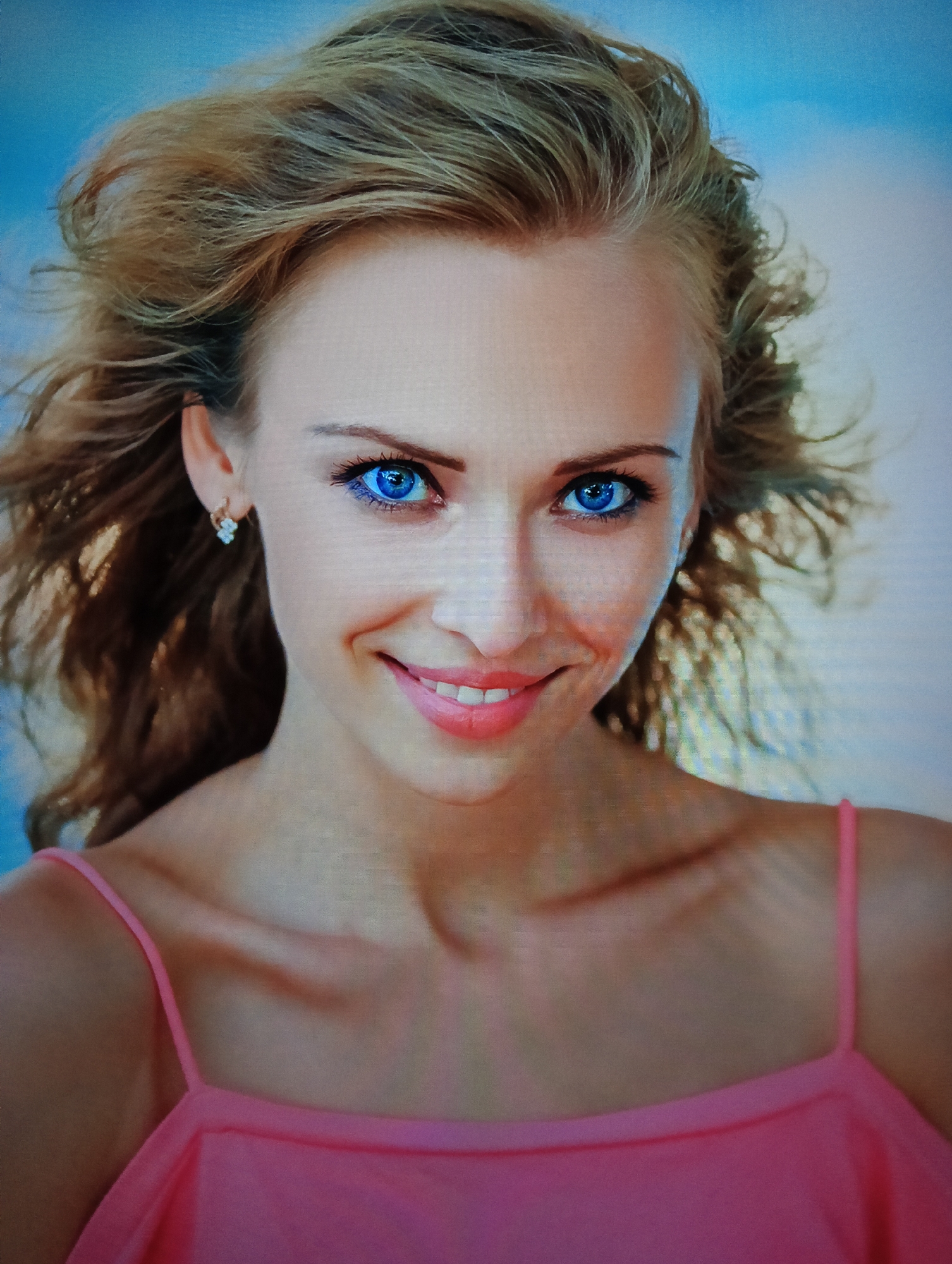 A close-up portrait of a smiling fair-skinned woman with blonde hair and striking blue eyes. She is wearing a pink top with spaghetti straps. An earring is visible in her left ear. The background is a muted blend of blue and white. The location in the city of Nha Trang Ward, Vietnam is indeterminable from the image provided.