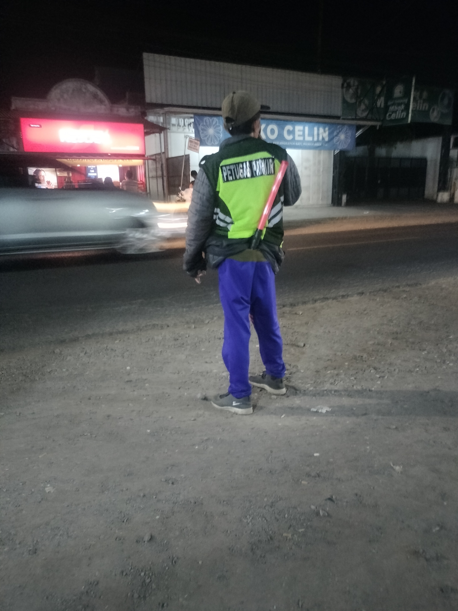 An outdoor, medium-shot image captures a person wearing a reflective vest and blue pants, standing on a gravel-strewn surface. The person is facing away from the camera, holding a bright orange and white striped signaling stick. The vest is a bright yellow-green with a black collar and shoulders, and the back of the vest has text that appears to read "PETUGAS PARKIR." The person is wearing a brown cap and dark gray sneakers. In the background, a building with a large red illuminated sign is visible, along with a smaller sign that says "KO CELIN." The scene is dimly lit, suggesting it is nighttime or dusk. The presence of vehicles and the context of a parking attendant suggest this location is likely a parking area in Srono, Indonesia, possibly near a business establishment.