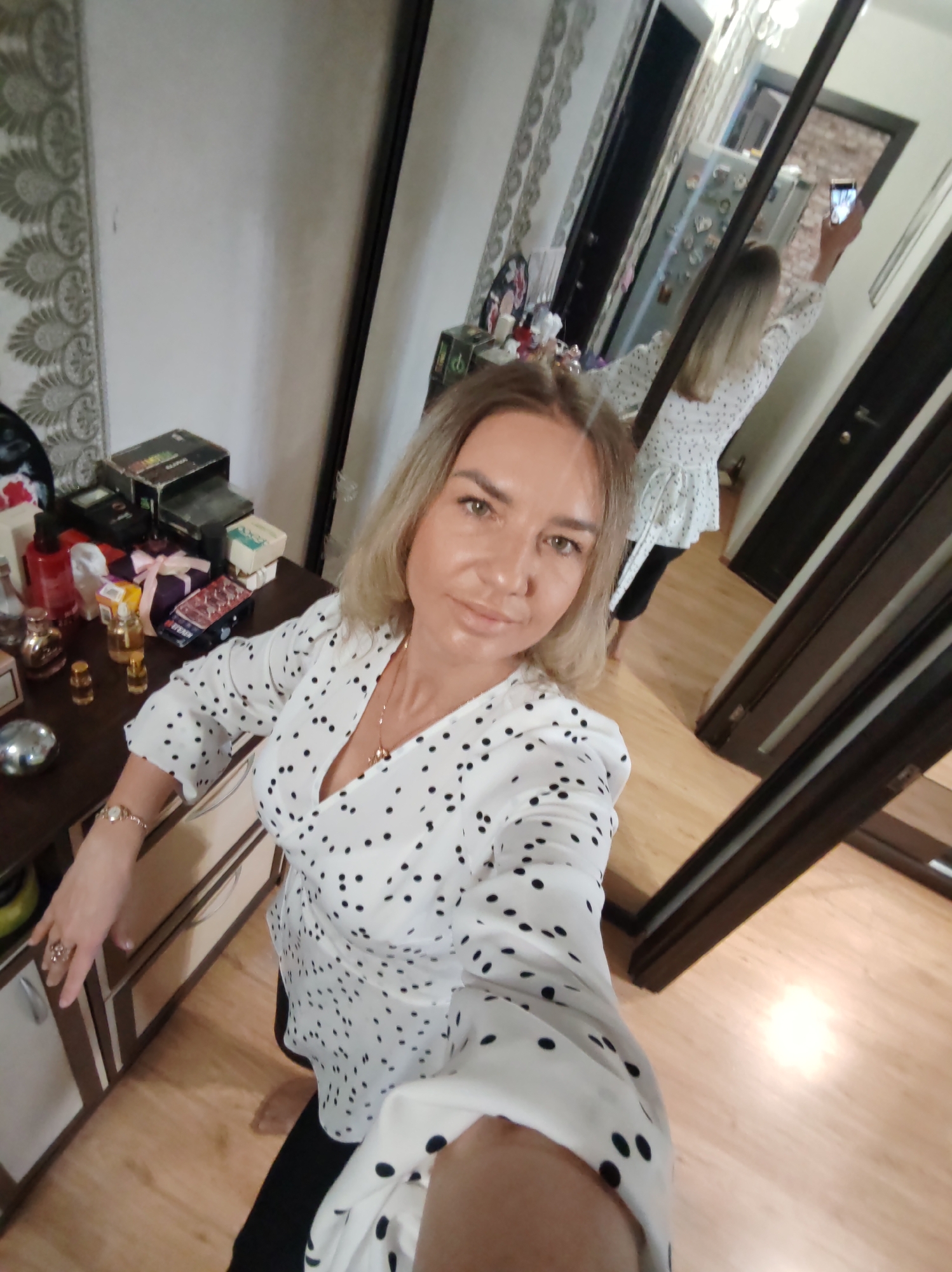 An indoor mirror selfie captures a woman in a casual setting, likely a bedroom or dressing area. In the foreground, the woman faces the camera, wearing a white blouse with black polka dots and black pants. She has blonde hair, a fair complexion, and a soft, neutral expression. A ring is visible on her right hand, and a watch is on her left wrist.
Her reflection is prominently displayed in a large mirror to the right, showing her from behind as she holds a smartphone aloft to take the photograph. The room features light-colored wooden flooring and patterned wallpaper on the left. To the left of the woman, a dresser or cabinet holds an assortment of personal items, including several perfume bottles, decorative bowls, and various boxes. Text on some boxes includes "ГАРАНТИЯ" (GUARANTEE) and "СССР" (USSR) in Cyrillic, suggesting vintage items, and "PARFUME" on another box. The mirror's reflection also reveals glimpses of the room behind the woman, including a door and a cabinet or refrigerator adorned with various magnets or decorations. The overall scene depicts a relaxed moment of self-documentation in a personal space.