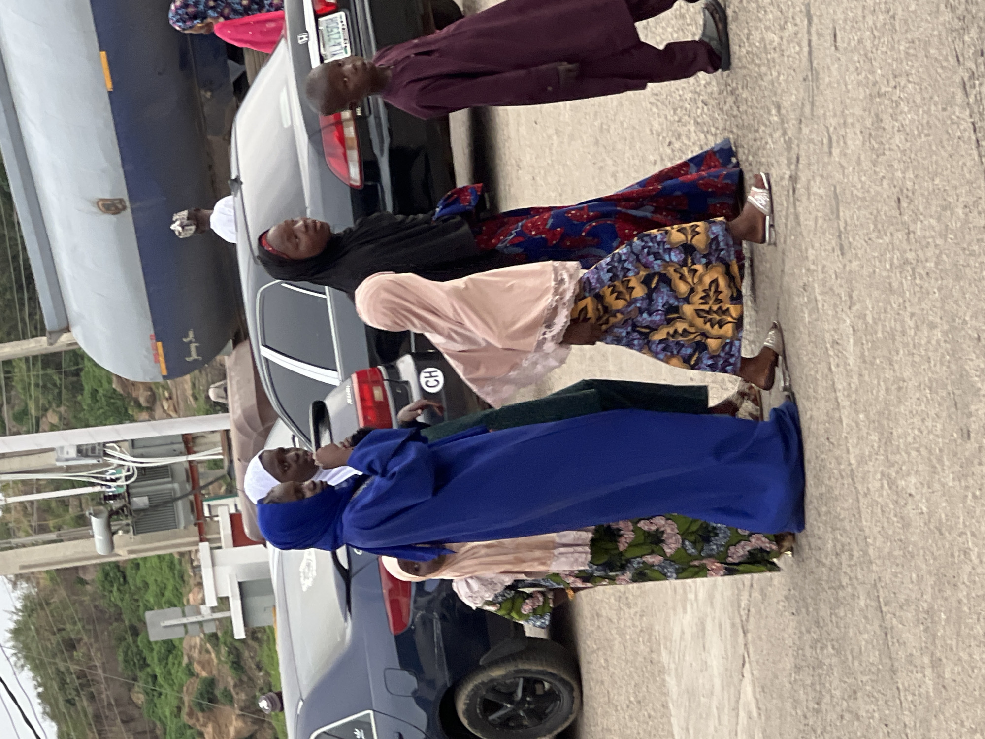A group of people, predominantly women wearing colorful robes and headscarves, are standing near cars on a paved street in Gura Top, Nigeria. A boy in a maroon robe is also visible. A large tanker truck is parked nearby. Electrical equipment and green vegetation can be seen in the background.