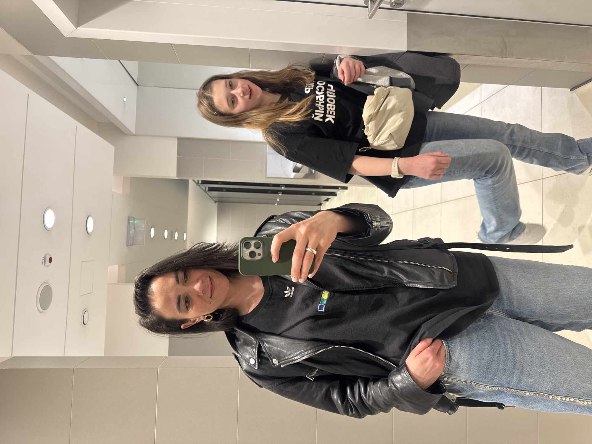 Two women are taking a selfie in a public restroom. The woman in the foreground is wearing a black leather jacket, a black Adidas t-shirt, and blue jeans. She is holding a green smartphone. The woman in the background is wearing a black t-shirt with white Cyrillic text, blue jeans, and a cream-colored bag. The restroom has white walls, bright lighting, and metal fixtures. The location may be a public restroom in a shopping mall, business, or entertainment venue in Belgrade, Serbia.