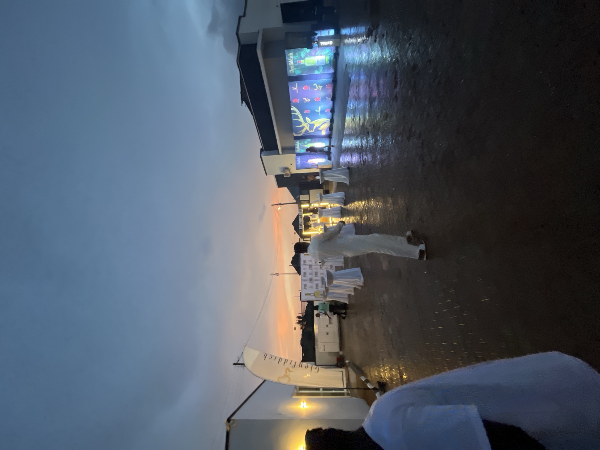 The image depicts an outdoor event at dusk in Bar Gada, Nigeria. A man in a white suit stands in the foreground, facing away from the camera, on a wet, reflective surface that appears to be asphalt. In the background, a large banner displays the Glenfiddich logo. Several high-top tables draped in white cloths are set up, some with people standing around them. Further in the background, a wall is illuminated with a vibrant light display featuring a deer graphic and bottles, also branded with Glenfiddich. The sky shows the orange hues of sunset. The overall scene suggests a promotional or social gathering.