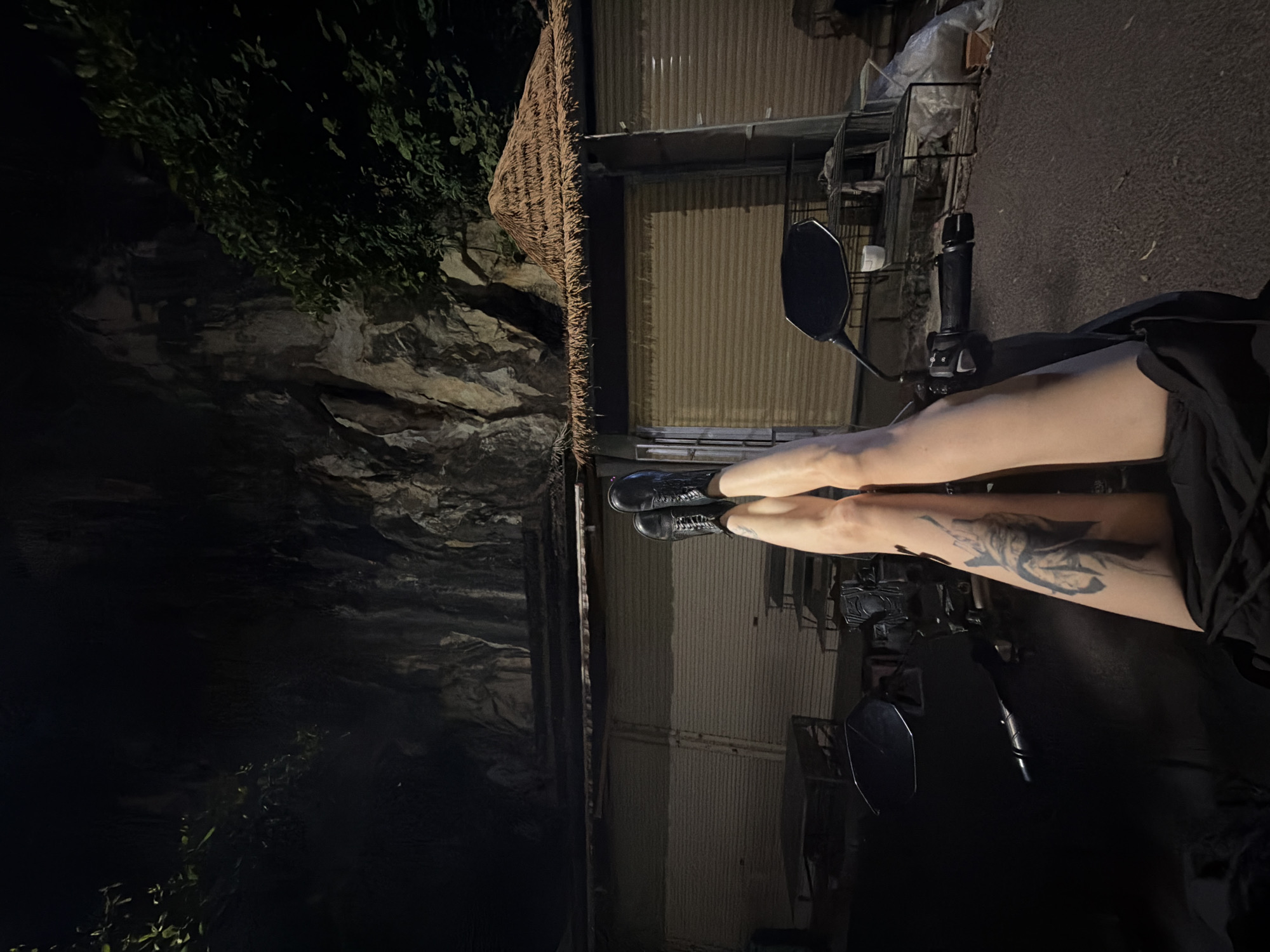 A nighttime view shows the legs of an individual in black boots, extended in front of a parked motorbike. The person is wearing black clothing. On one leg is a tattoo. The bike's handlebars and mirror are visible. In the background, there is a corrugated metal building topped with a thatched roof, which suggests a street-side establishment. Adjacent to the building is a natural rock wall with vegetation, illuminated by artificial lighting. This combination of natural elements and urban infrastructure suggests a location within Đà Nẵng, Vietnam, where such features are common. The scene suggests the person is stationary and resting on the parked vehicle, observing the surroundings.