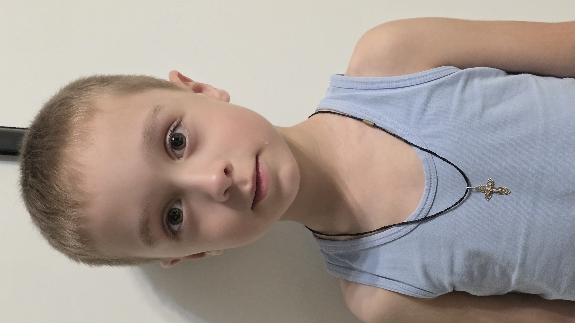 A young boy with a light complexion, short blonde hair, and brown eyes is seen wearing a light blue tank top and a black cord necklace with a silver cross pendant. The boy is facing the camera at an angle. The background is a plain white wall. The location is unknown.