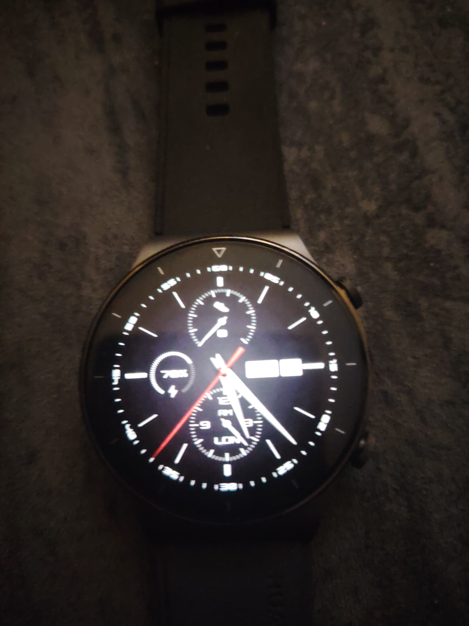 A black smartwatch with a circular display and a black textured strap is depicted on a dark, textured fabric surface within Huntingdonshire, United Kingdom. The scene is dimly lit, with the watch face emitting illumination. The watch face displays an analog interface with white hour and minute hands indicating approximately 11:55 or 12:55, and a red second hand pointing towards the lower left. A circular sub-dial on the left shows "78%" with a lightning bolt icon. A rectangular sub-display on the right indicates "MON" (Monday) and an obscured date. A third sub-dial at the bottom displays "AM" or "PM" and text possibly reading "LON" or "LOW" with an associated numerical value. A physical button is visible on the right side of the watch casing.