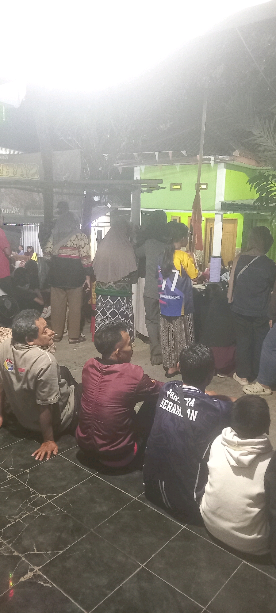 In the city of Argasoka, Indonesia, a group of people are gathered outdoors at night, likely for a social event or ceremony, under a bright light source that washes out much of the background. The foreground is dominated by a tiled floor. Several individuals are seated or kneeling on the floor, facing away from the camera.
In the lower left, a man wearing a khaki shirt with a logo is seated, leaning on his left hand. He appears to be smoking. To his right, another man in a maroon shirt is seated. Further to the right, a person in a navy blue t-shirt with white lettering is seated, and a person in a white hooded sweatshirt is also seated.
In the midground, several other individuals are standing. A woman in a patterned skirt and a headscarf is visible. A young person in a yellow and blue jersey with the number "77" is standing. Other figures in the background are less distinct due to the lighting and distance. The environment appears to be a courtyard or open area, with a glimpse of a brightly painted green building in the background, suggesting it might be a residential or community space. Small triangular flags are strung across an overhead structure, adding to the festive or ceremonial atmosphere.